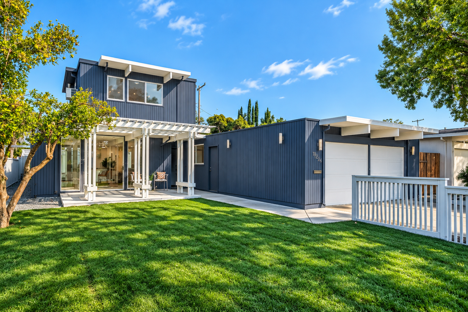 Modern two-story house with blue siding, large glass doors, and a well-maintained front lawn. The house features a white pergola over the porch and has a white fence on one side. The sky is clear and blue with a few clouds, and there are green trees around the property.