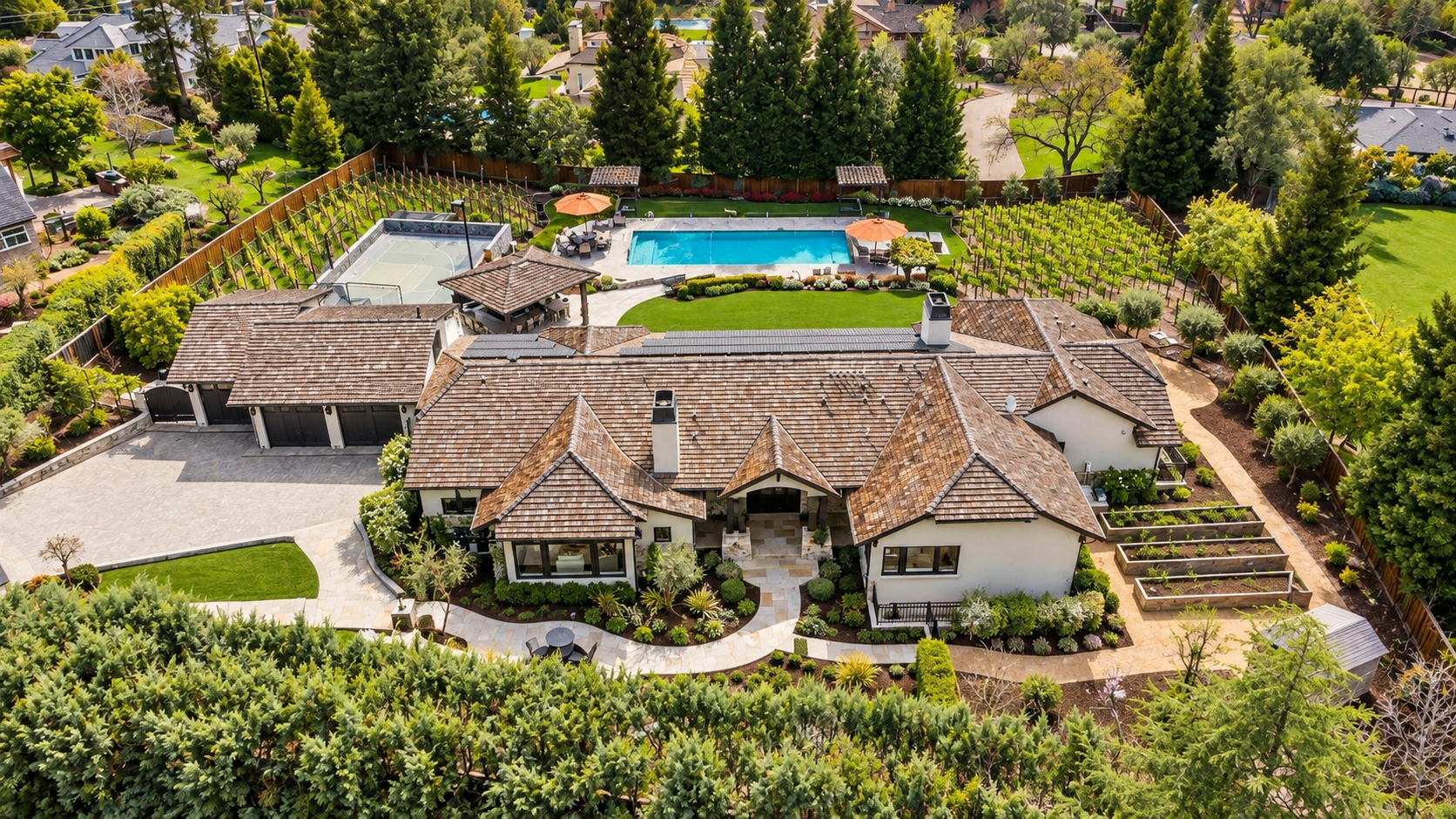 Aerial view of a large residential backyard with a house, swimming pool, landscaped garden, patio area with umbrellas, tennis court, and surrounding trees and lawn.