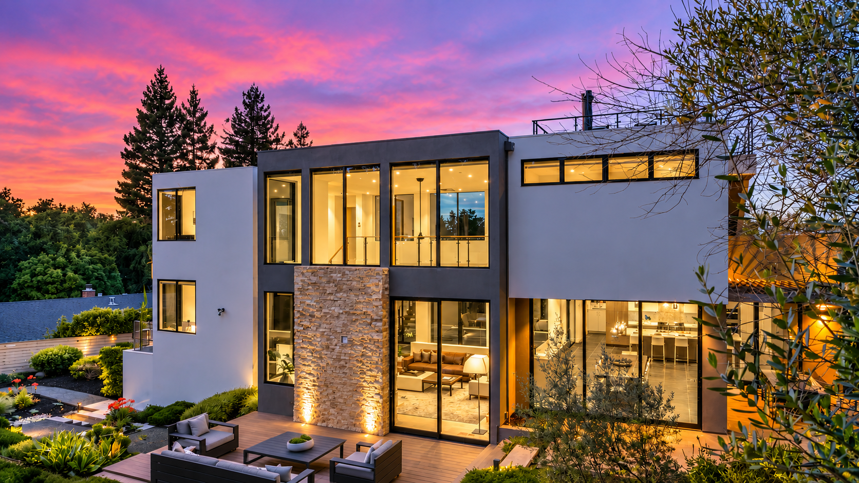 Modern multi-story house with large glass windows during sunset, surrounded by greenery and outdoor seating area.