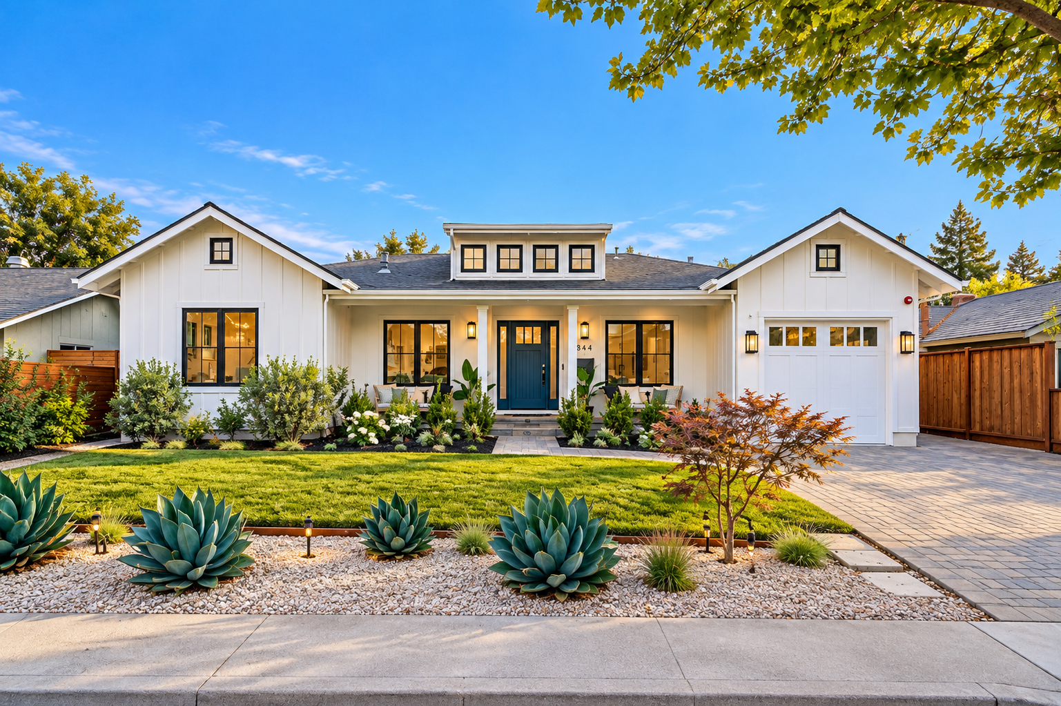 Front view of a modern white house with black window frames, a blue front door, and a landscaped yard with succulents, shrubs, and a small tree, under a bright blue sky.