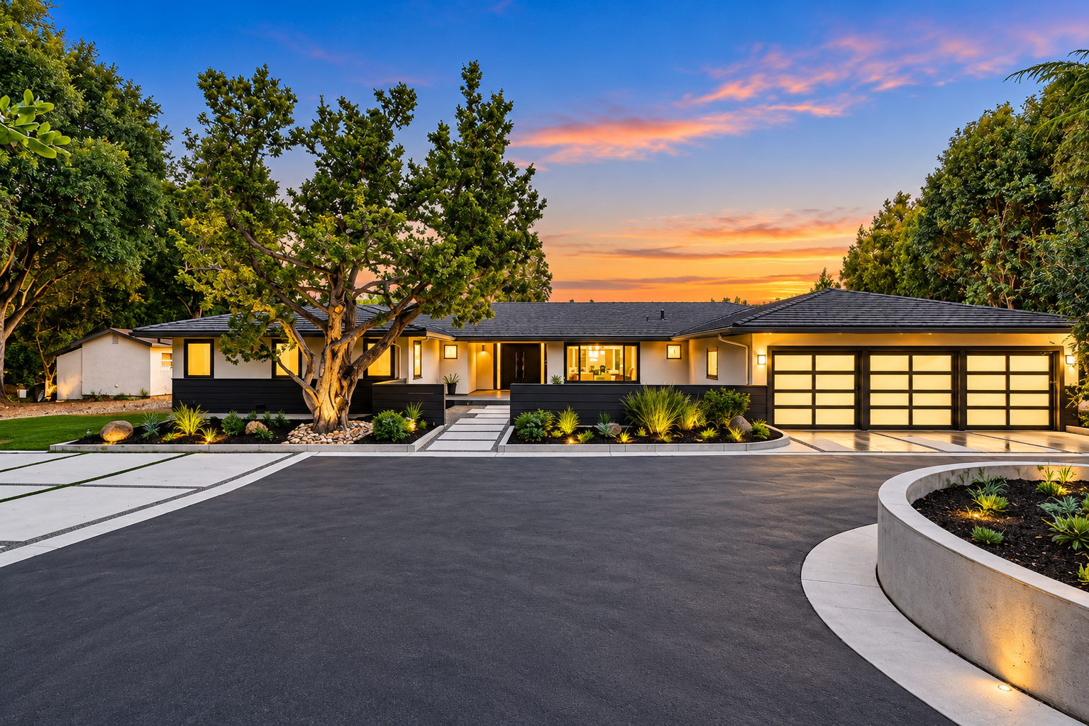 Modern house with a large driveway, illuminated exterior lighting, and landscaped front yard with trees and shrubs during sunset.