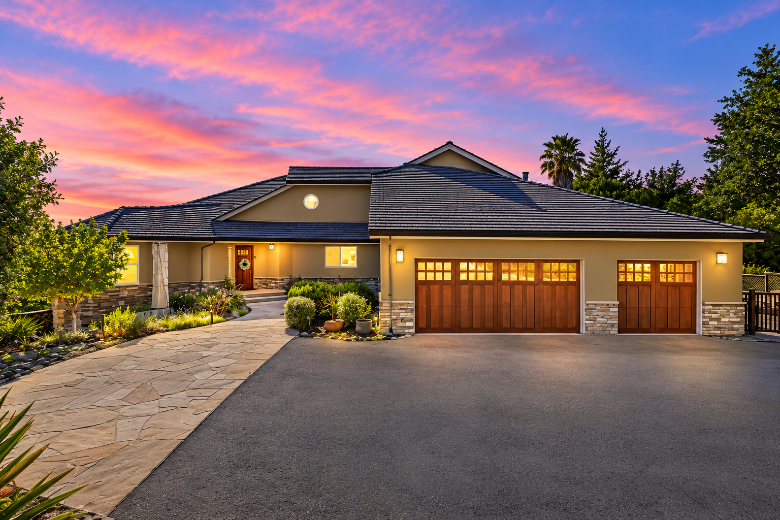 A modern house with stone and wood accents on the garage and front door, surrounded by well-maintained landscaping, under a colorful sunset sky.