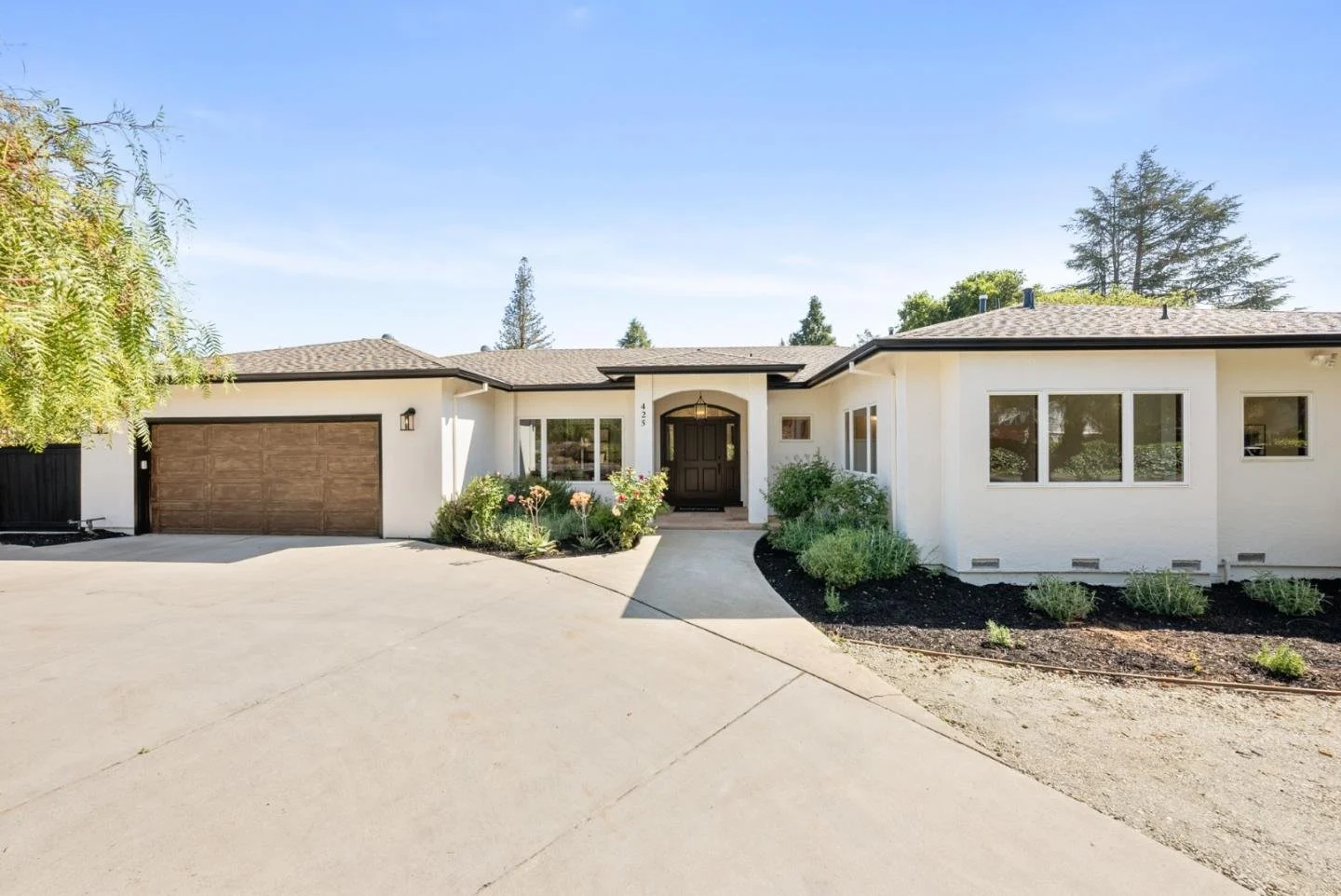 Single-story white house with brown garage door, front door, and landscaped garden with shrubs and flowers, under a blue sky.