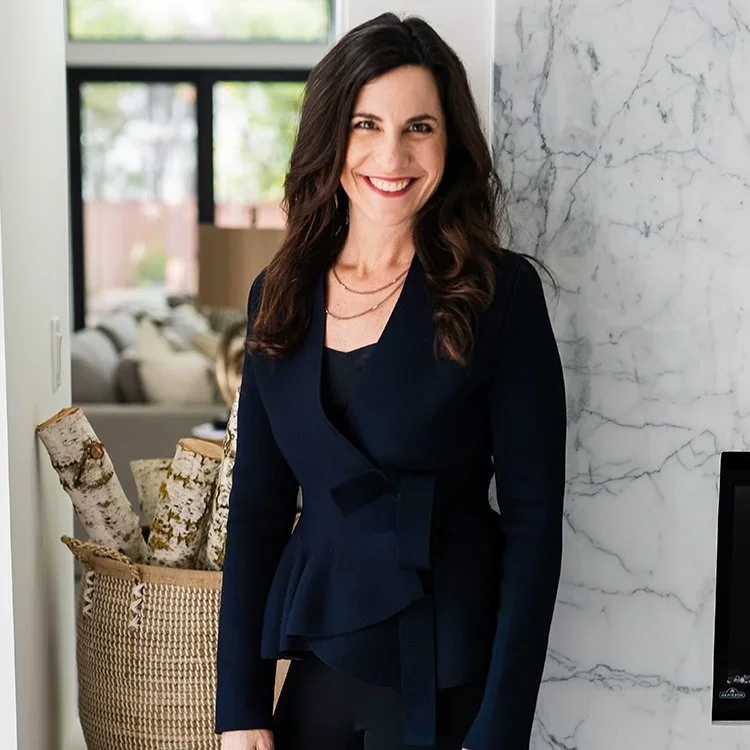 A woman with long dark hair and a bright smile standing indoors near a marble wall, wearing a navy blazer or dress, with a living room visible in the background.