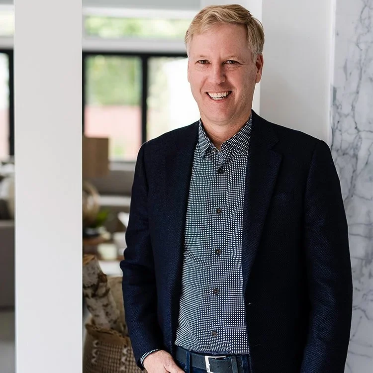 A smiling man in a navy blazer and patterned shirt standing inside a modern home or office with large windows.