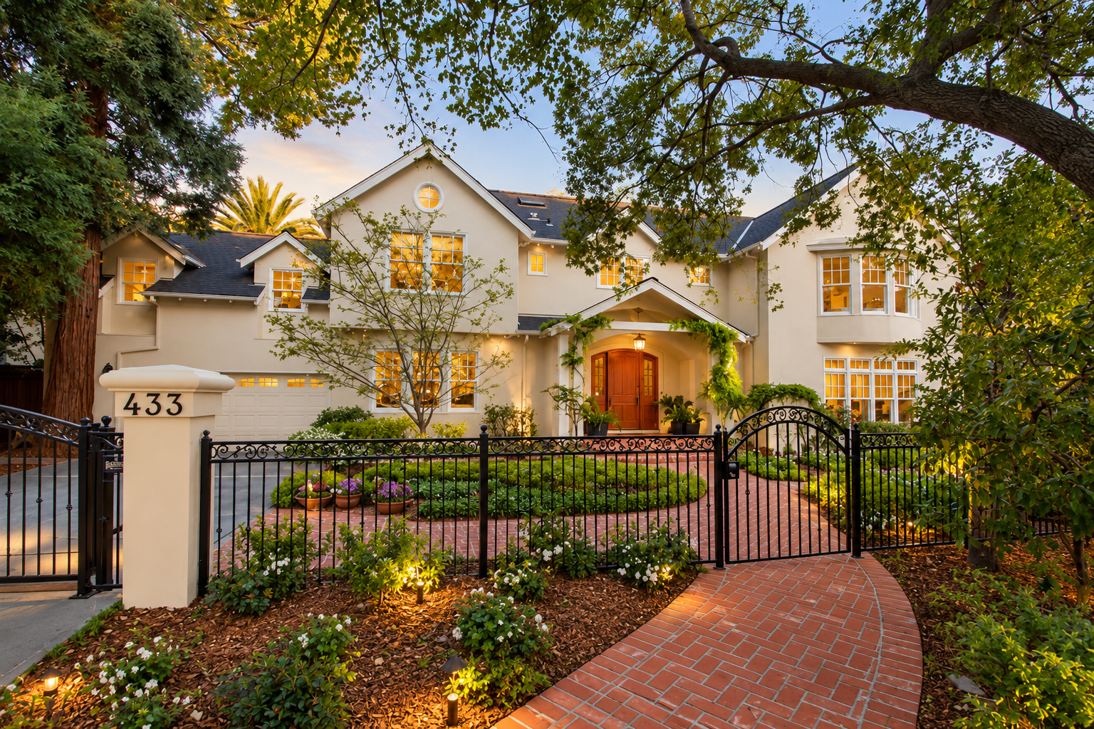 Front yard of a large, white house with a brick pathway leading to a wooden front door, surrounded by a black metal fence, trees, and landscaped garden with flowers, during sunset.