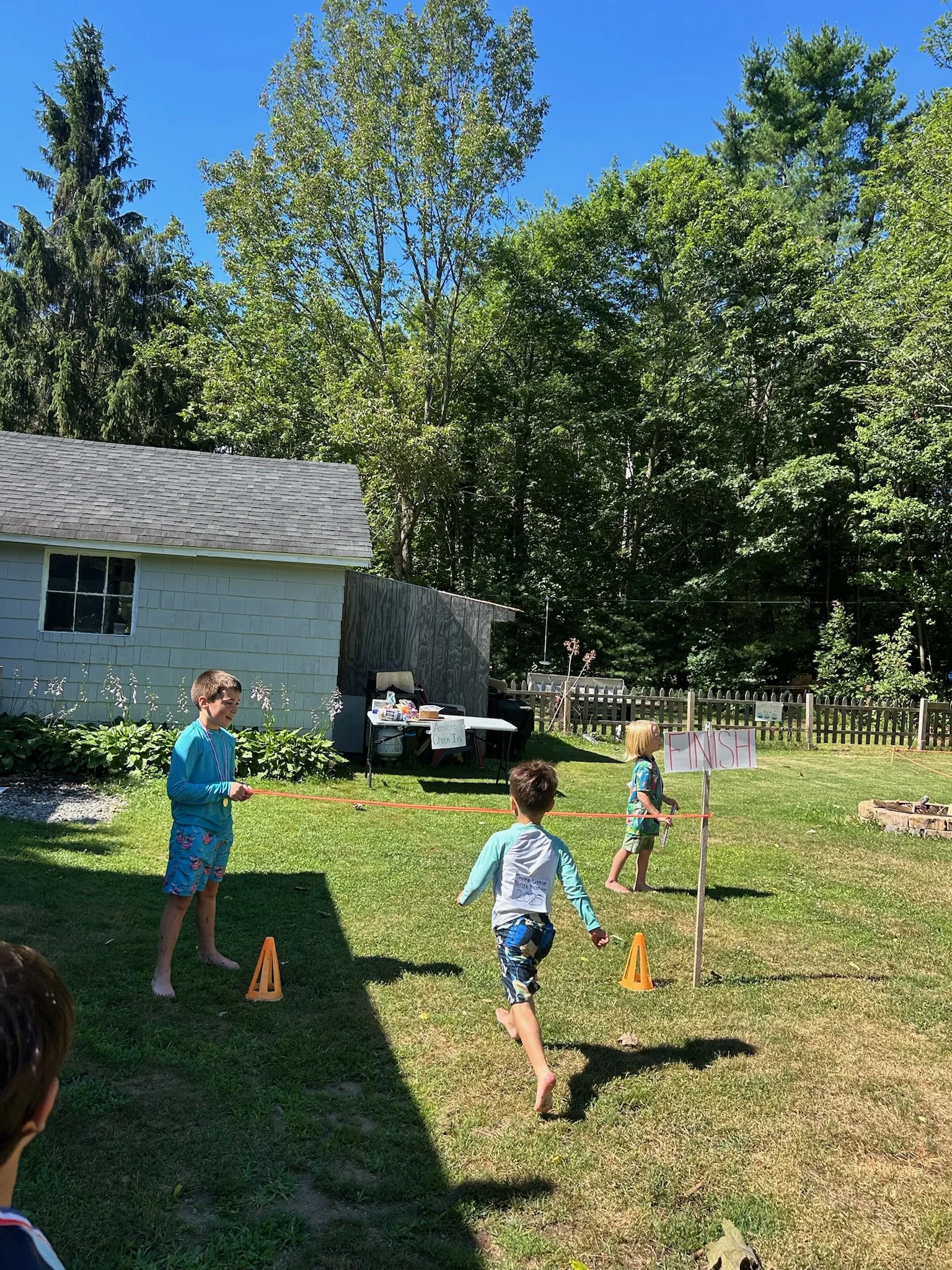 Children participating in an outdoor race or game in a backyard with a sign reading 'Finish'. There are cones marking the track, a table with items, and a house with trees in the background under a clear blue sky.
