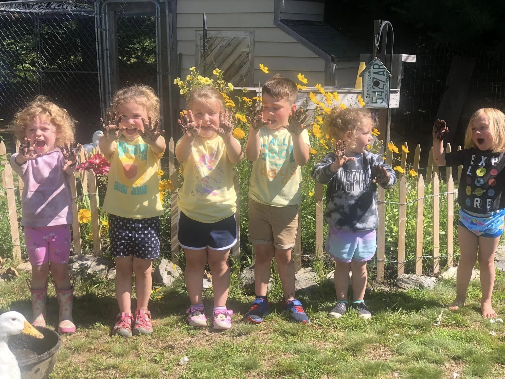 Six young children standing in a row outdoors, displaying dirt-covered hands, smiling and looking at the camera. They are behind a small wooden fence with yellow flowers, in a garden area with a house and a fenced yard in the background.