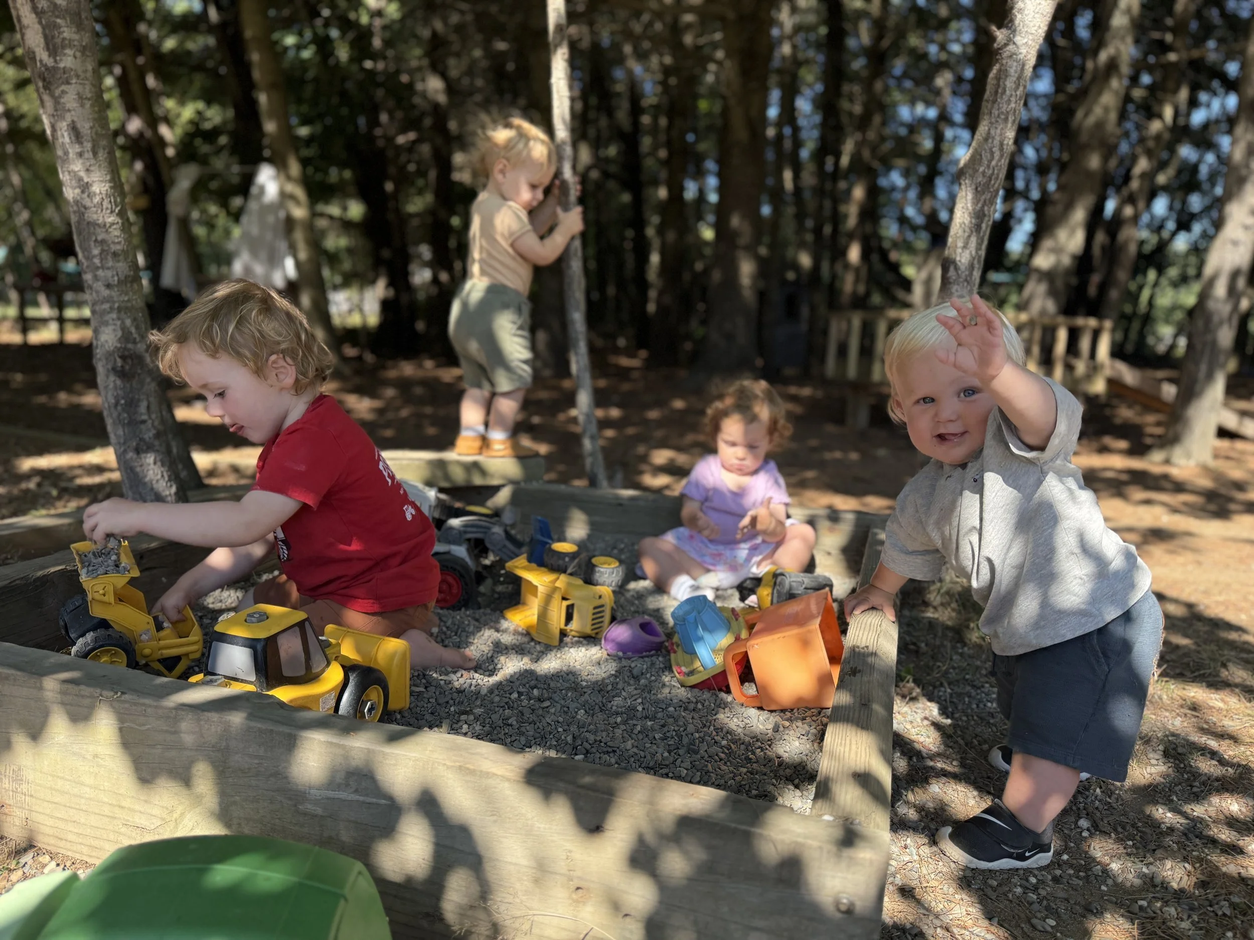 Children playing in a sandbox at a park, surrounded by trees with sunlight filtering through.