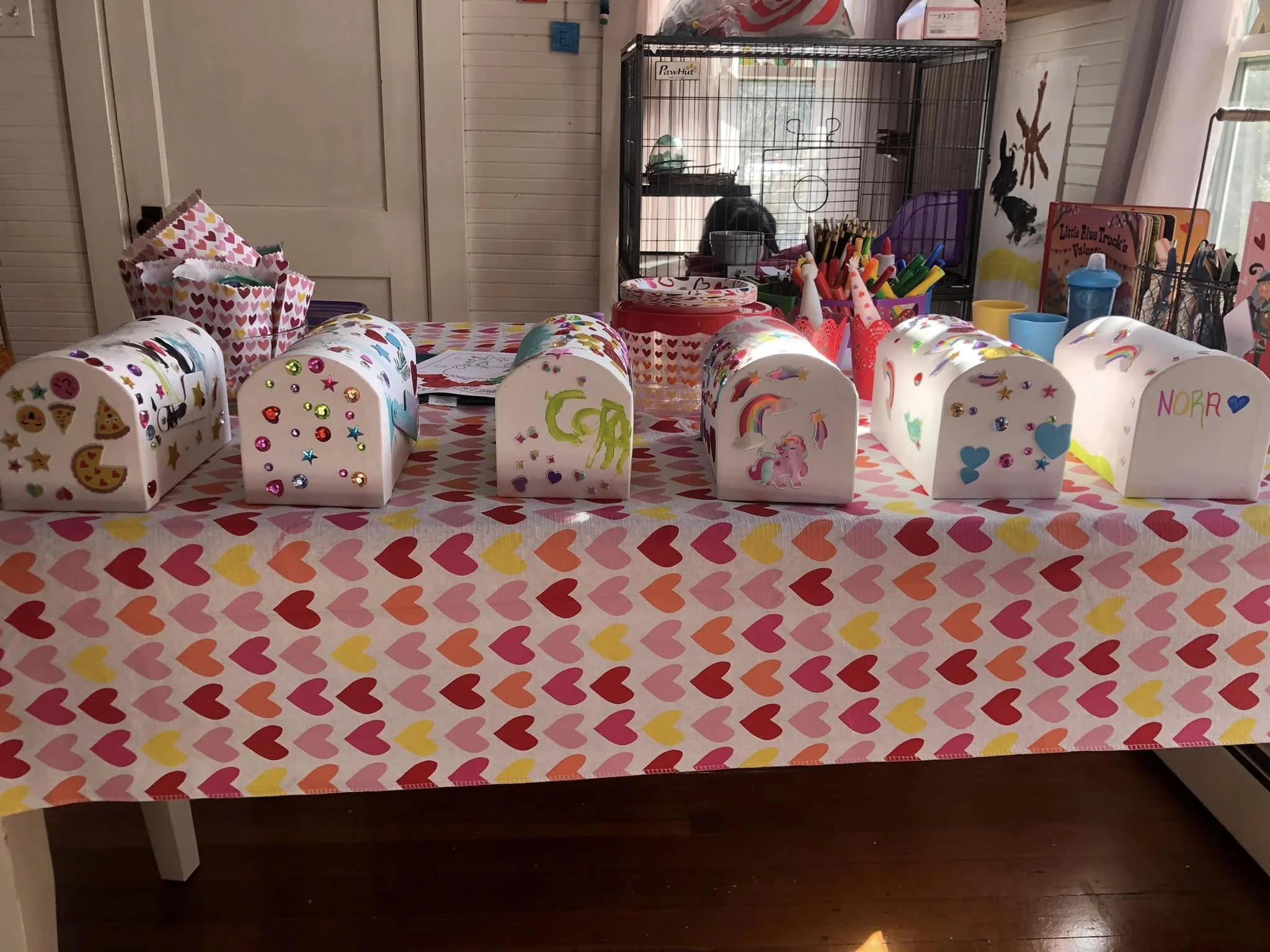 Valentine's Day craft table decorated with heart-themed items, including small gift boxes, cupcakes, and colorful stickers, with a pet cage in the background.