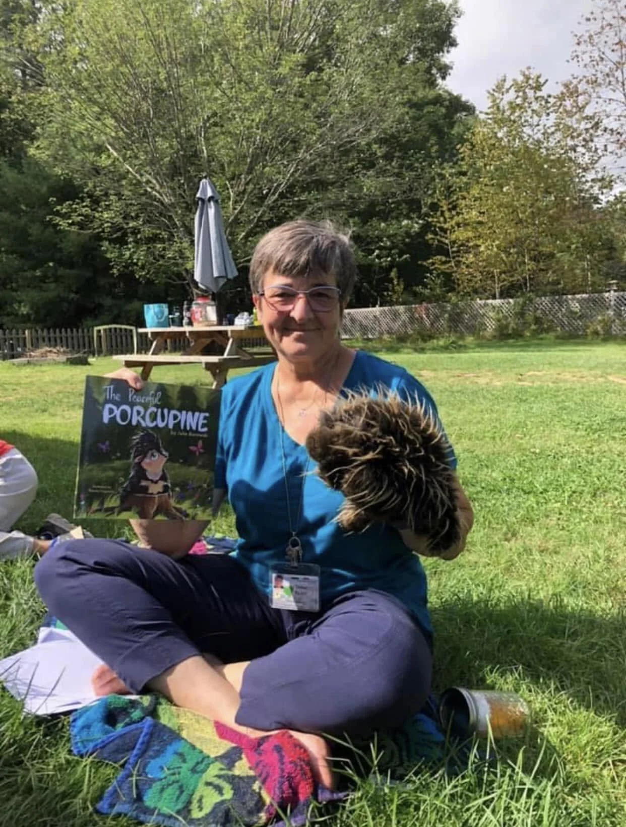 A woman sitting cross-legged on the grass outdoors, holding a cute porcupine in her hand, with a book titled 'The People of Porcupine' beside her, and a picnic scene in the background with a picnic table, an umbrella, and trees.