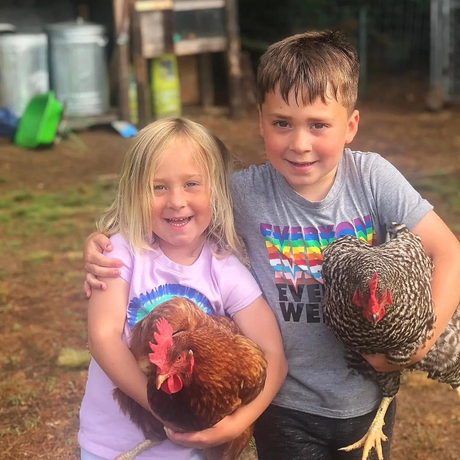 Two children, a girl and a boy, holding chickens, smiling at the camera outdoors with farm equipment in the background.