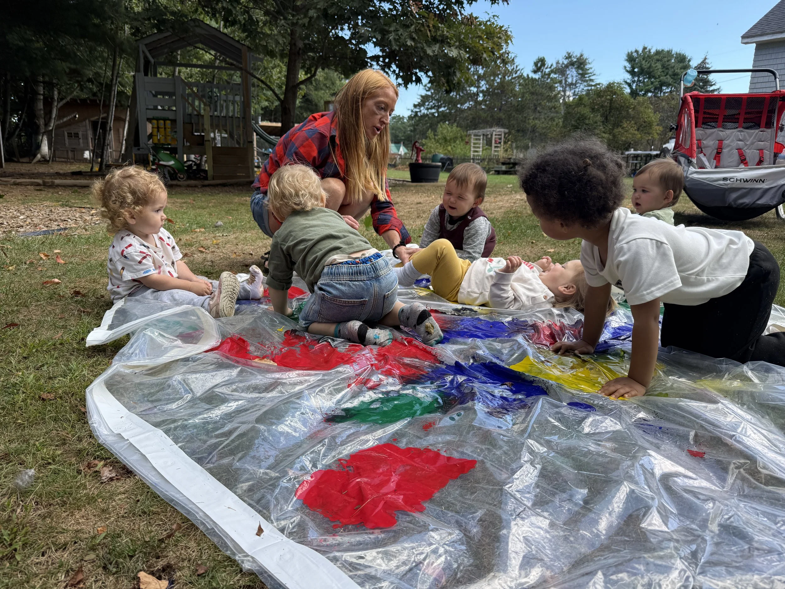 A woman with red hair and a red plaid shirt surrounded by five small children on a grassy outdoor area, engaging in a painting activity with colors on a large plastic sheet, with trees and a playground in the background.
