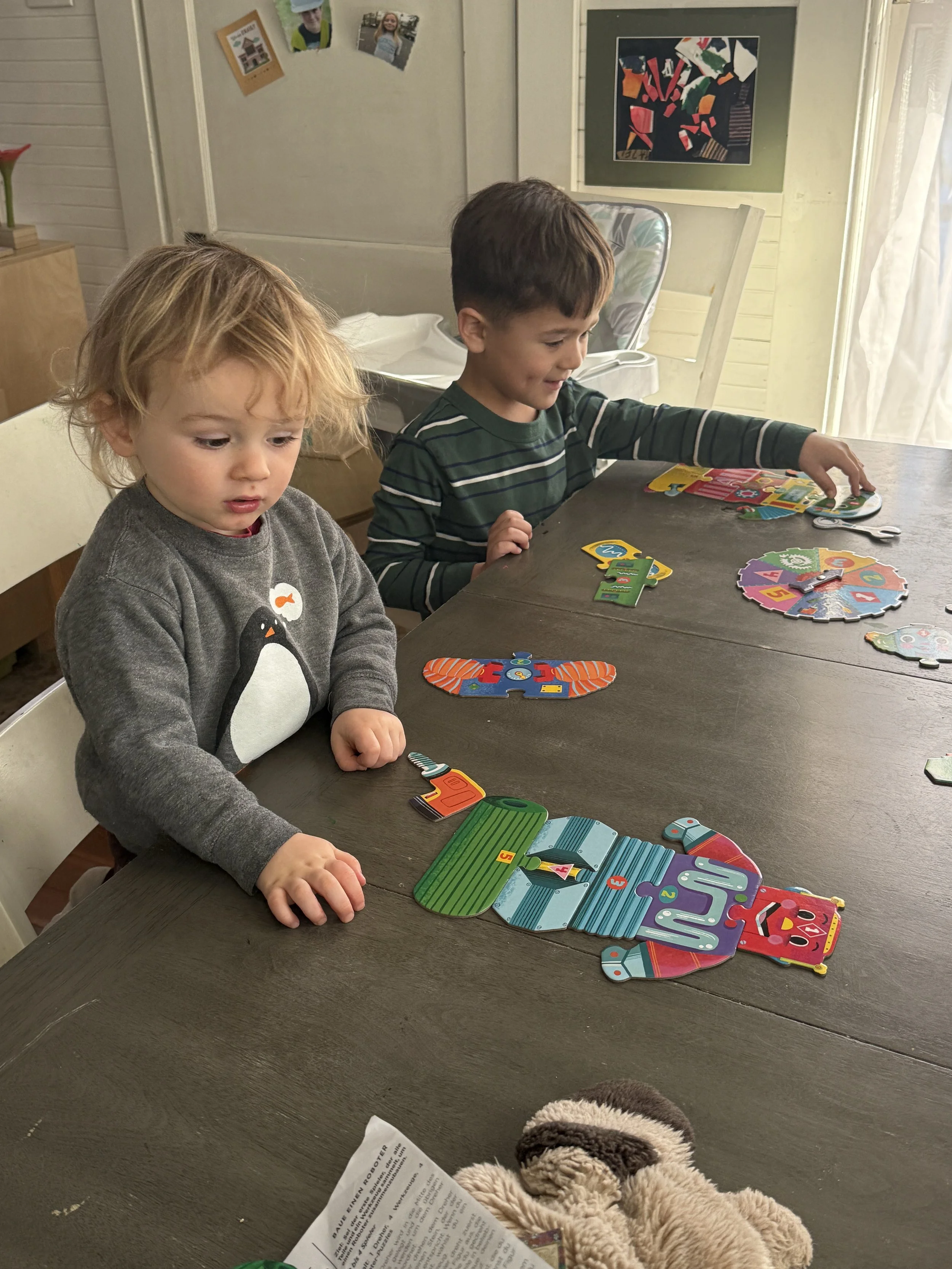 Two young children sitting at a dark wooden table playing with colorful jigsaw puzzles. The child on the left has blonde hair and wears a gray sweater with a penguin on it. The child on the right has brown hair and wears a green and white striped shi