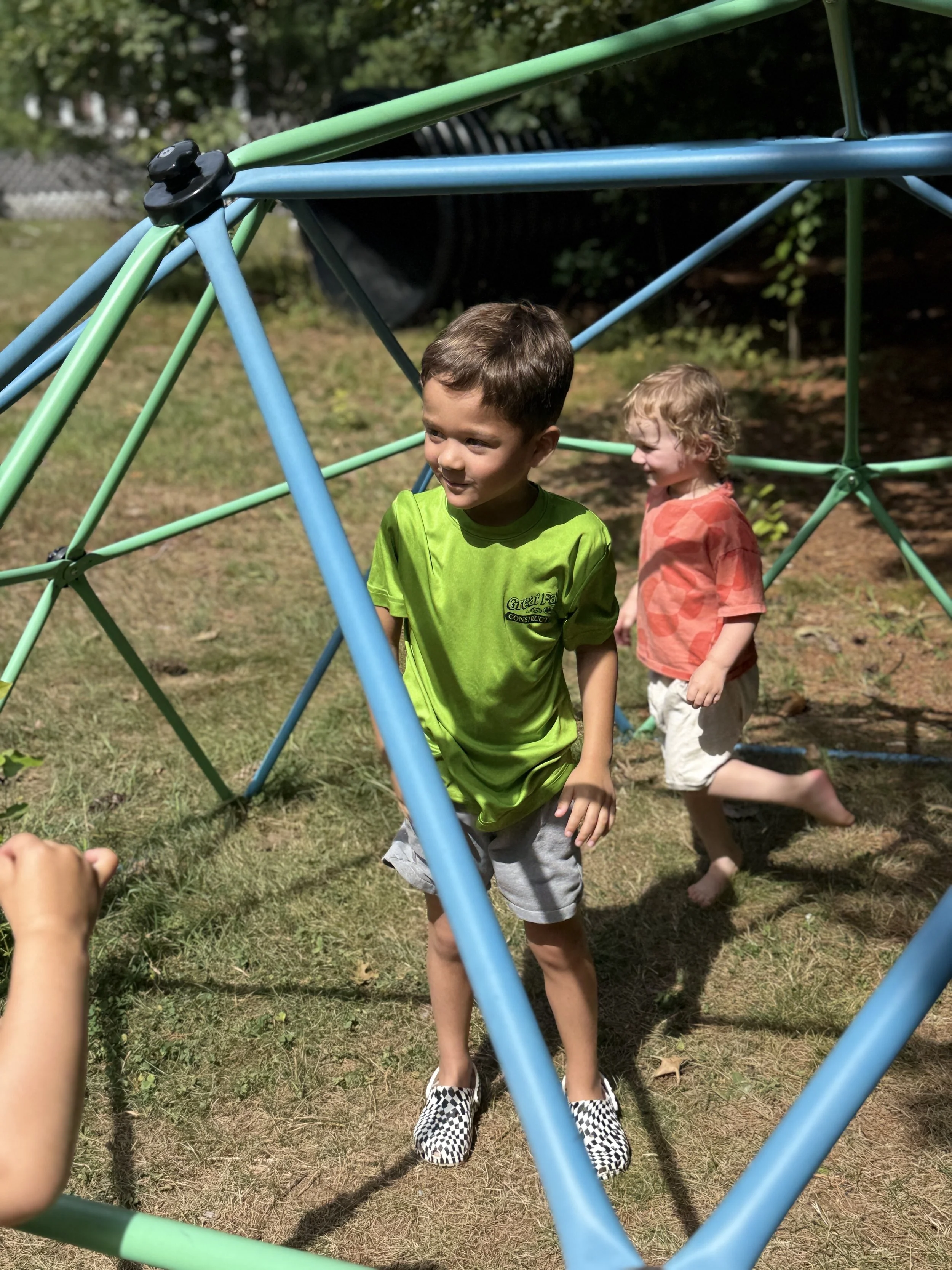 Two young boys playing outside on a grassy area near a metal jungle gym structure. One boy in a green shirt and checkered shoes is in the foreground, and another boy in a red shirt and beige shorts is behind him. The background has trees and a trash 