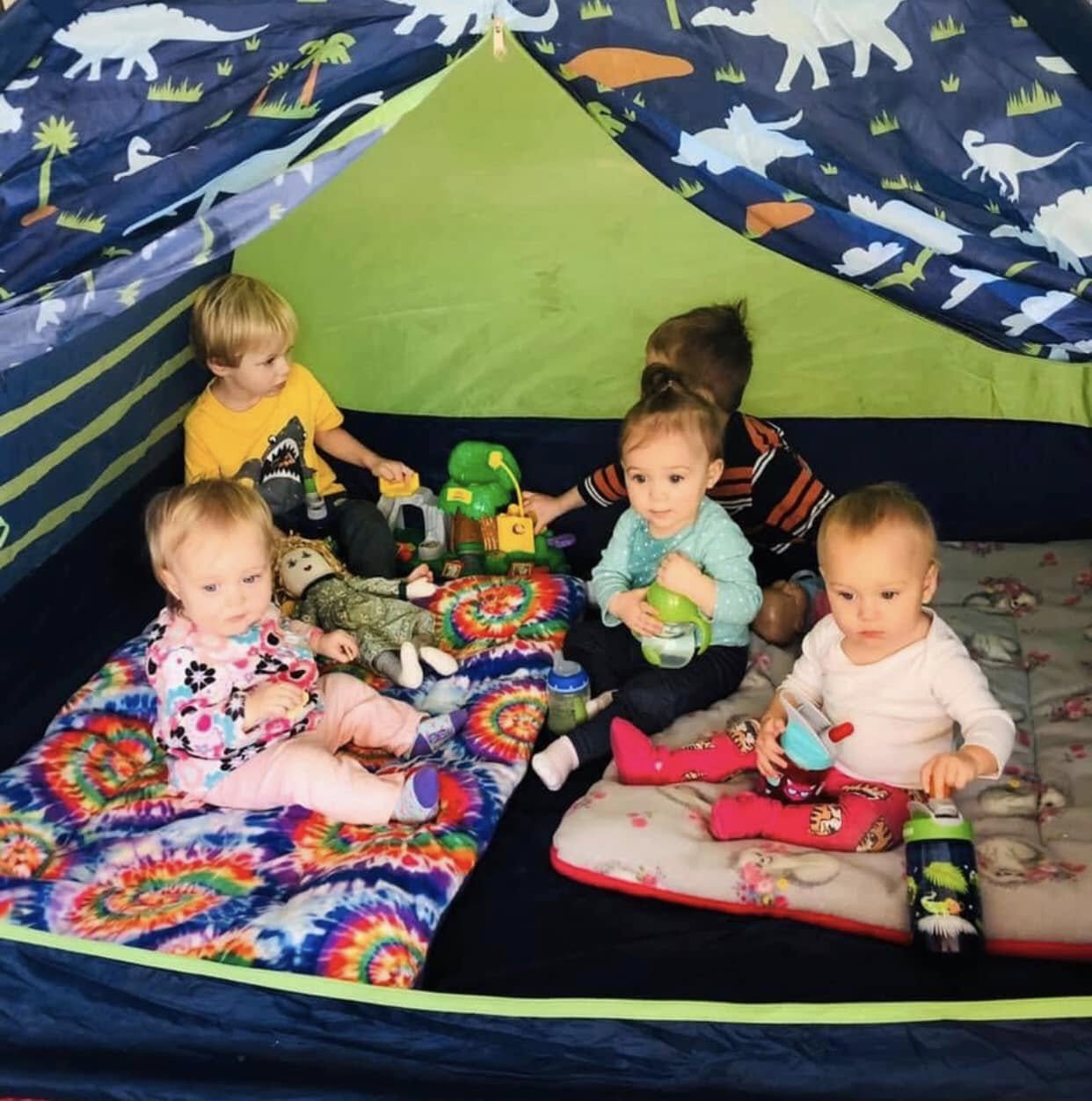 A group of young children sitting inside a colorful play tent with dinosaur and jungle print, surrounded by toys and blankets, some holding cups and bottles.