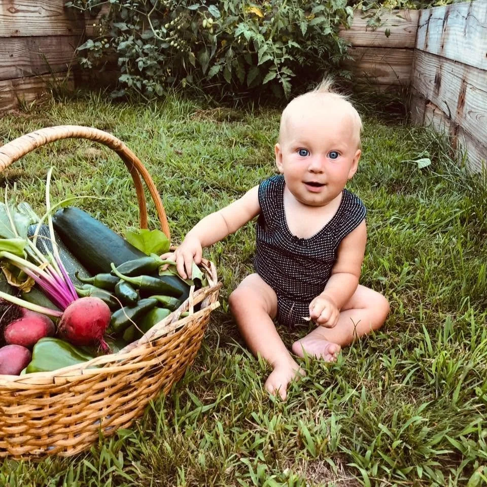 A baby with blonde hair and blue eyes sitting on grass in a garden, next to a basket of fresh vegetables including radishes, cucumbers, and green onions.