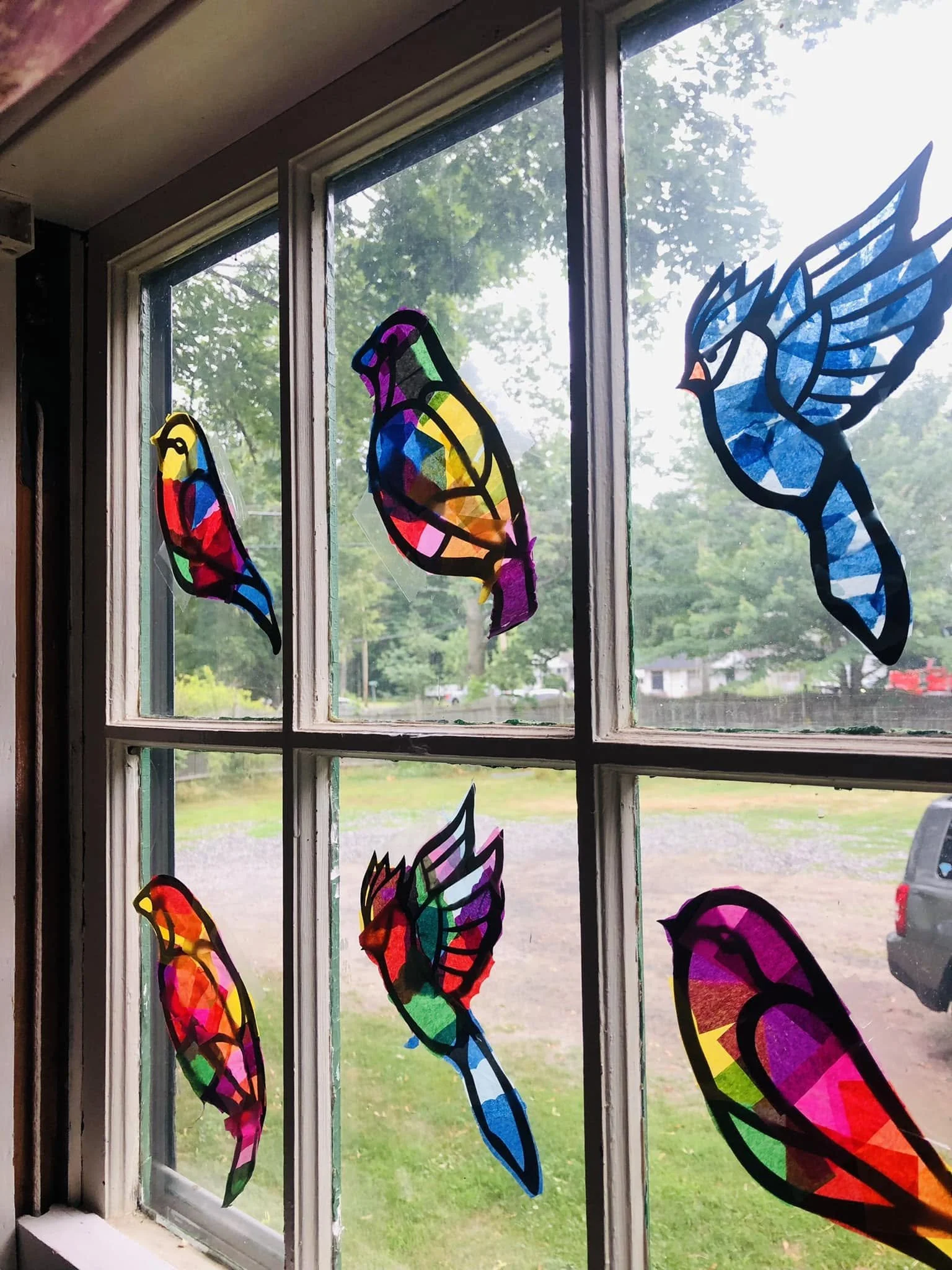 Colorful stained glass bird decorations on a window with a view of green trees and a lawn outside.