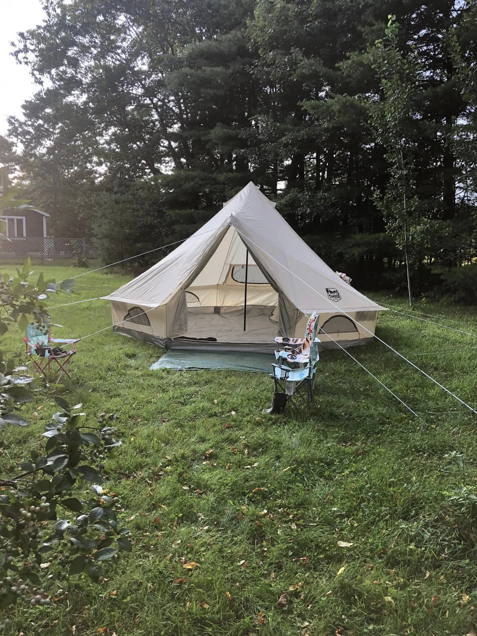 A beige canvas tent set up on a grassy yard with trees in the background. There are small chairs and a mat in front of the tent, and a house with a deck is visible to the left.