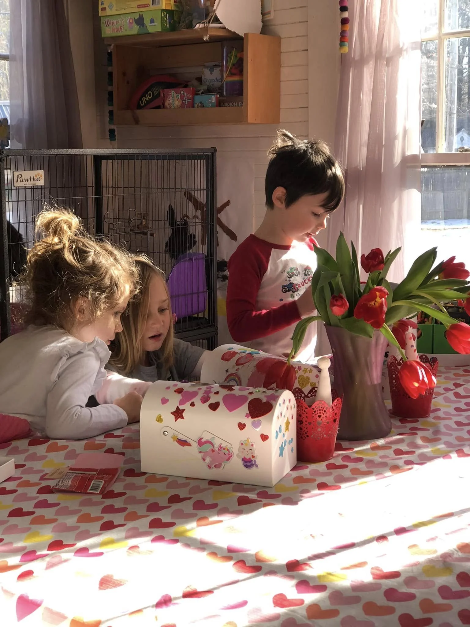 A birthday celebration with children gathered around a table with Valentine's Day themed decorations. There are three children visible, two girls and a boy, with the girl on the left having curly hair and the girl in the middle with straight hair. Th