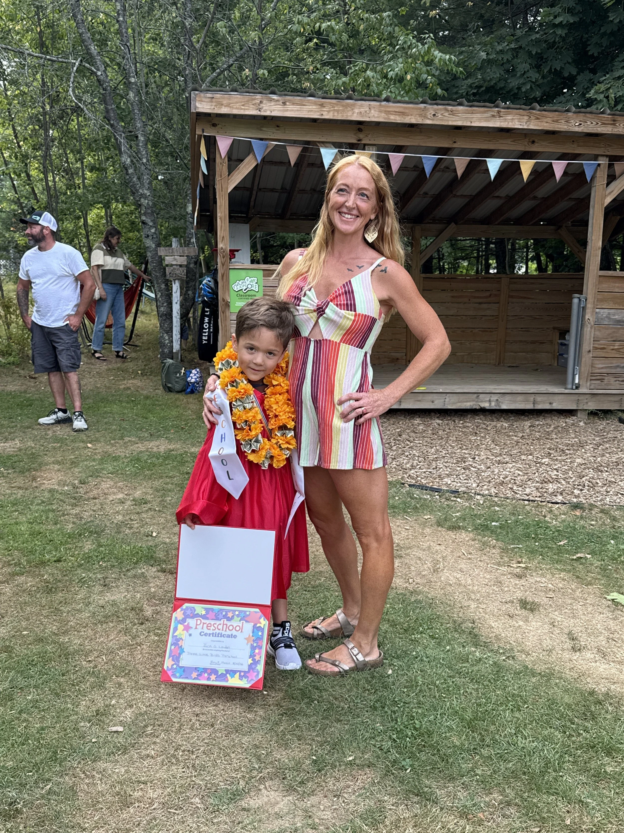 A woman and a young girl standing outdoors on grass with trees in the background. The woman is wearing a multicolored striped dress, sandals, and has red hair. The girl is wearing a red dress with a sash that reads 'Preschool,' a flower lei, and snea
