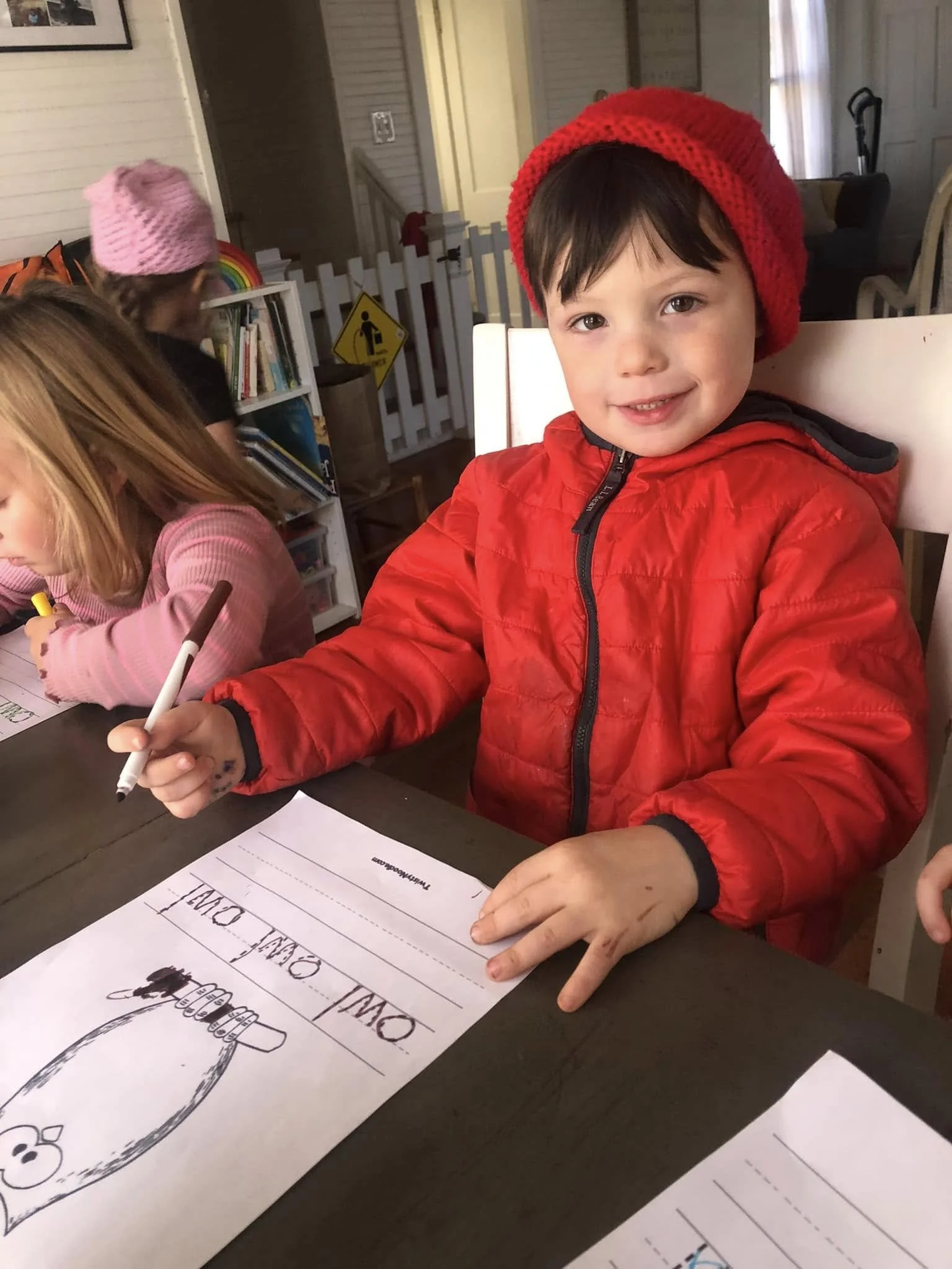 A young boy wearing a red jacket and red knitted hat sitting at a table, smiling at the camera while coloring on a sheet of paper with a marker. Two other children are in the background, one girl in a pink sweater and another girl with a pink knitted