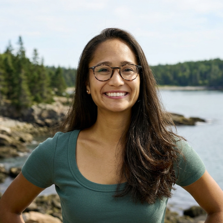 Smiling woman with glasses standing outdoors near a body of water with trees in the background.