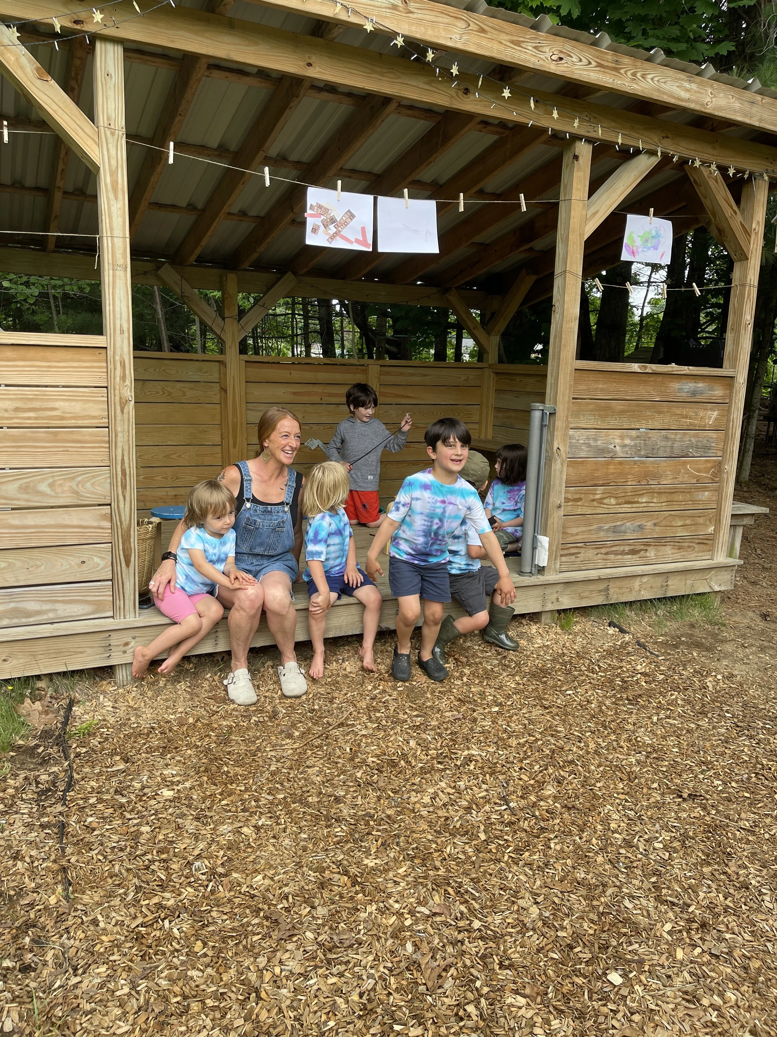 A group of children and a woman are at an outdoor wooden shelter with a sloped roof, surrounded by trees. The ground is covered with wood chips. Some children are sitting on the porch, and others are standing and moving around. There are drawings han