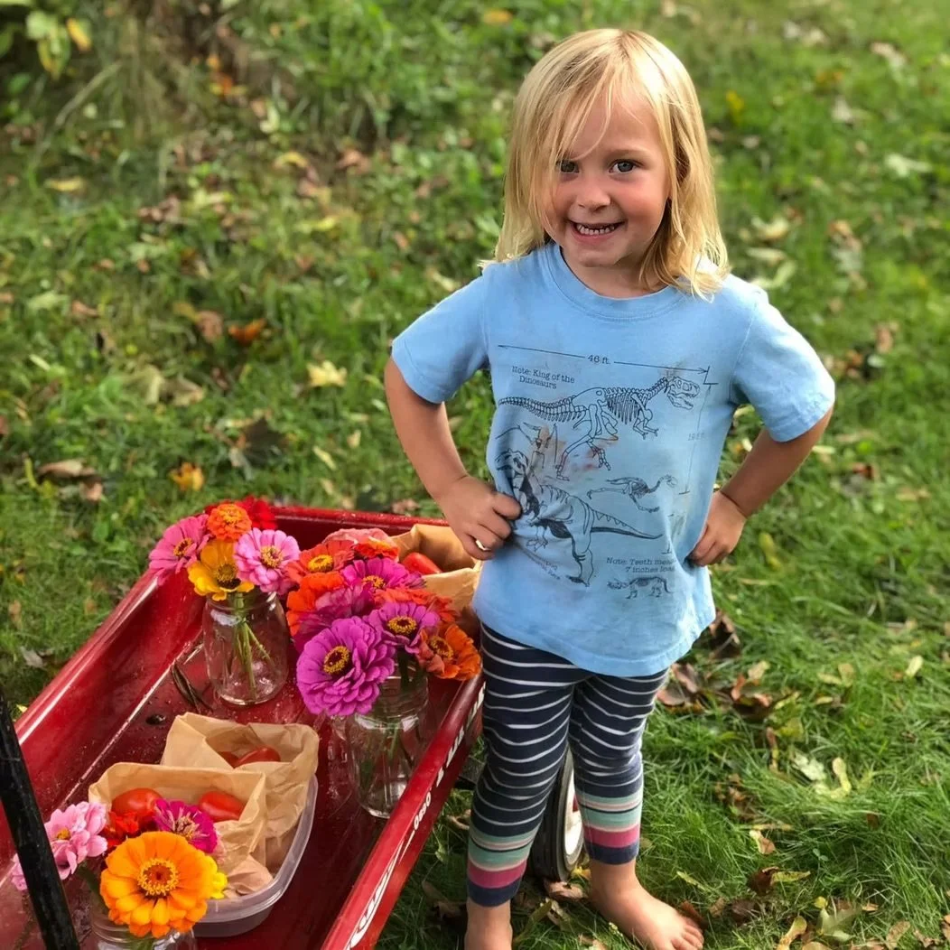 A young girl with blonde hair standing outdoors on grass, smiling with hands on her hips, next to a red wagon filled with vases of colorful flowers and paper bags containing tomatoes ready to sell at Soriel Farm Store in Scarborough, Maine.