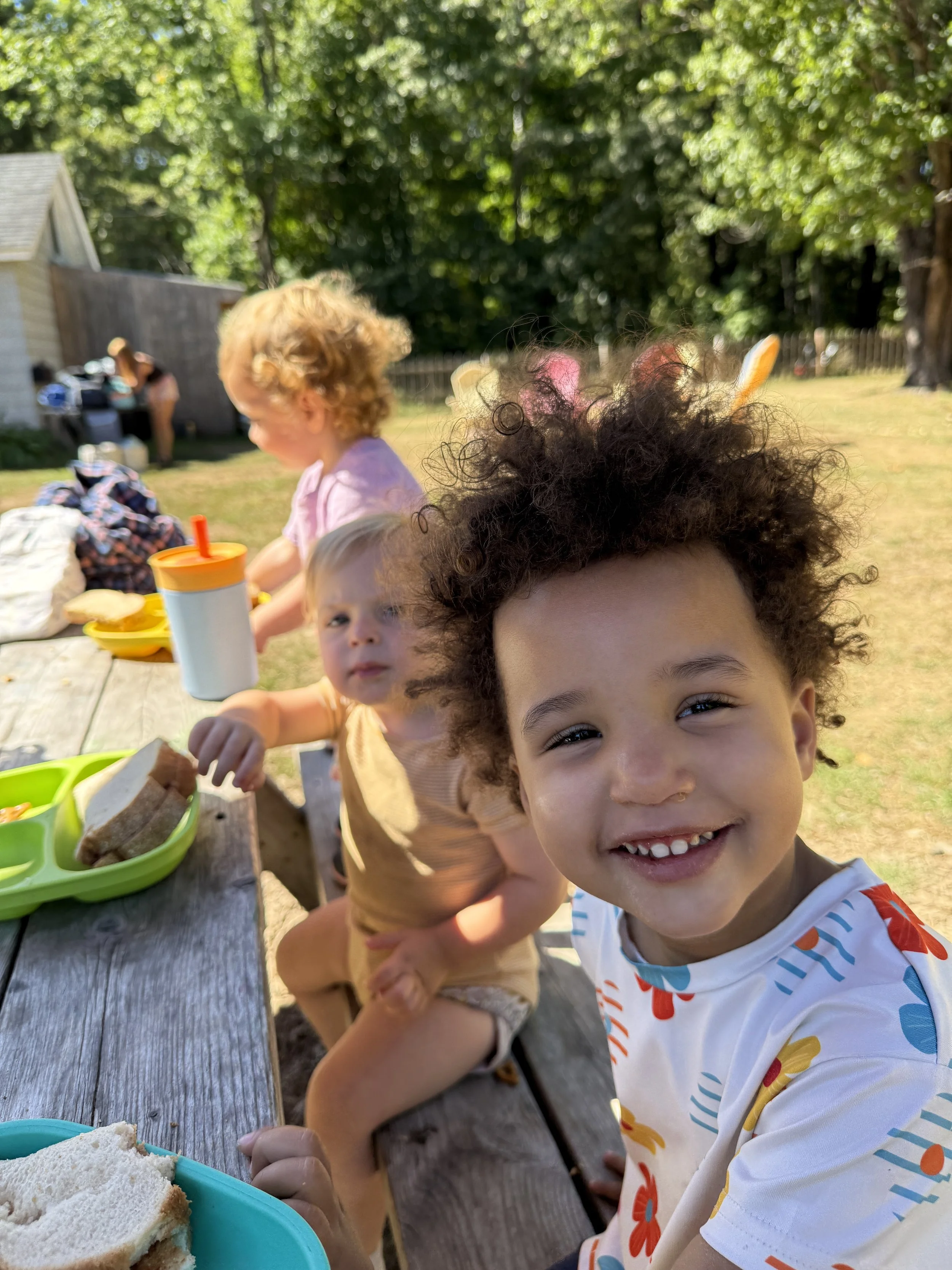 Smiling young boy with curly hair in a colorful T-shirt outdoors at a picnic table, with other children and a woman in the background, surrounded by green trees and a backyard fence.