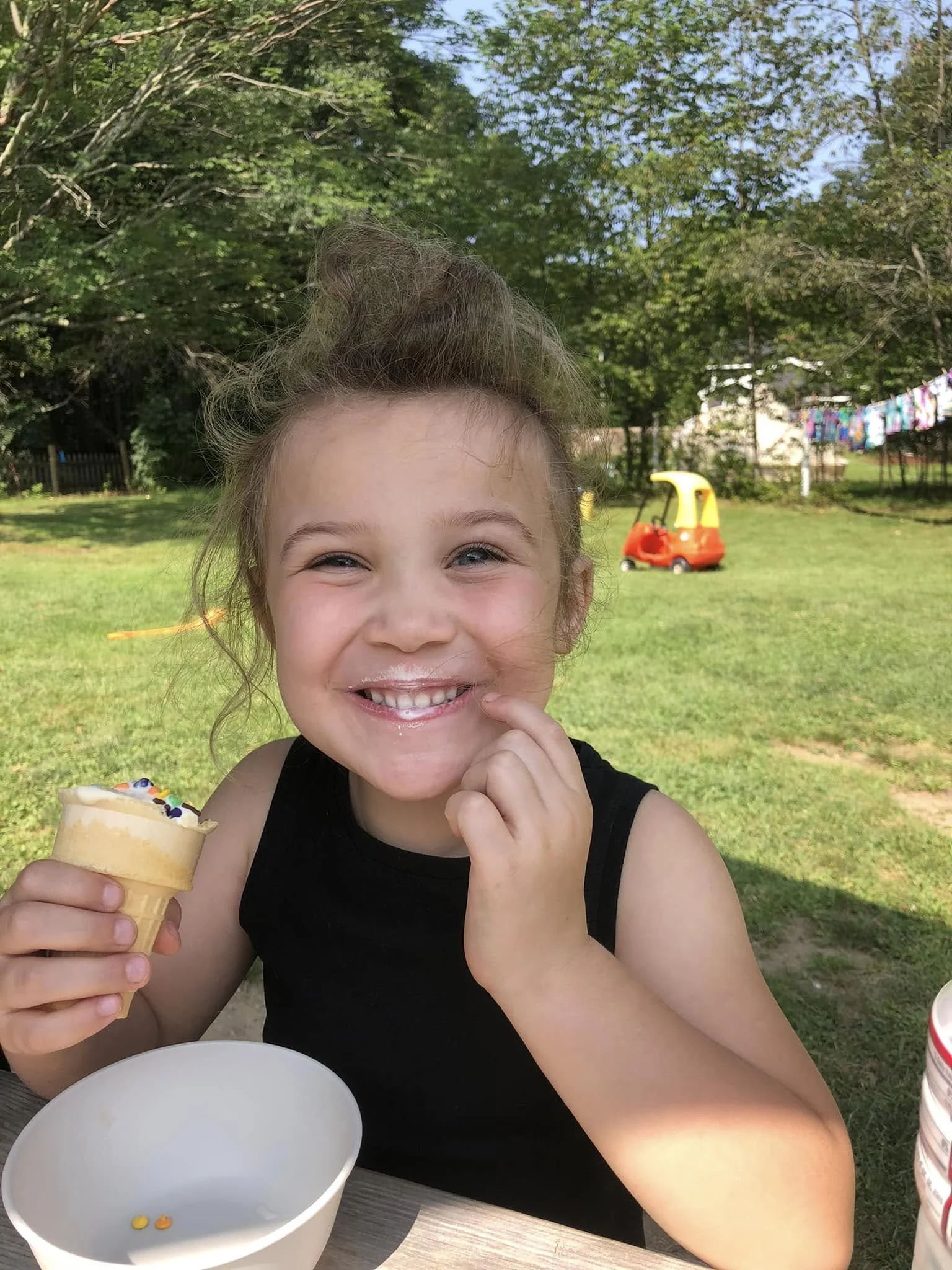 A young girl smiling while holding an ice cream cone outdoors in a backyard with trees, laundry hanging, and a small red and yellow toy car in the background.