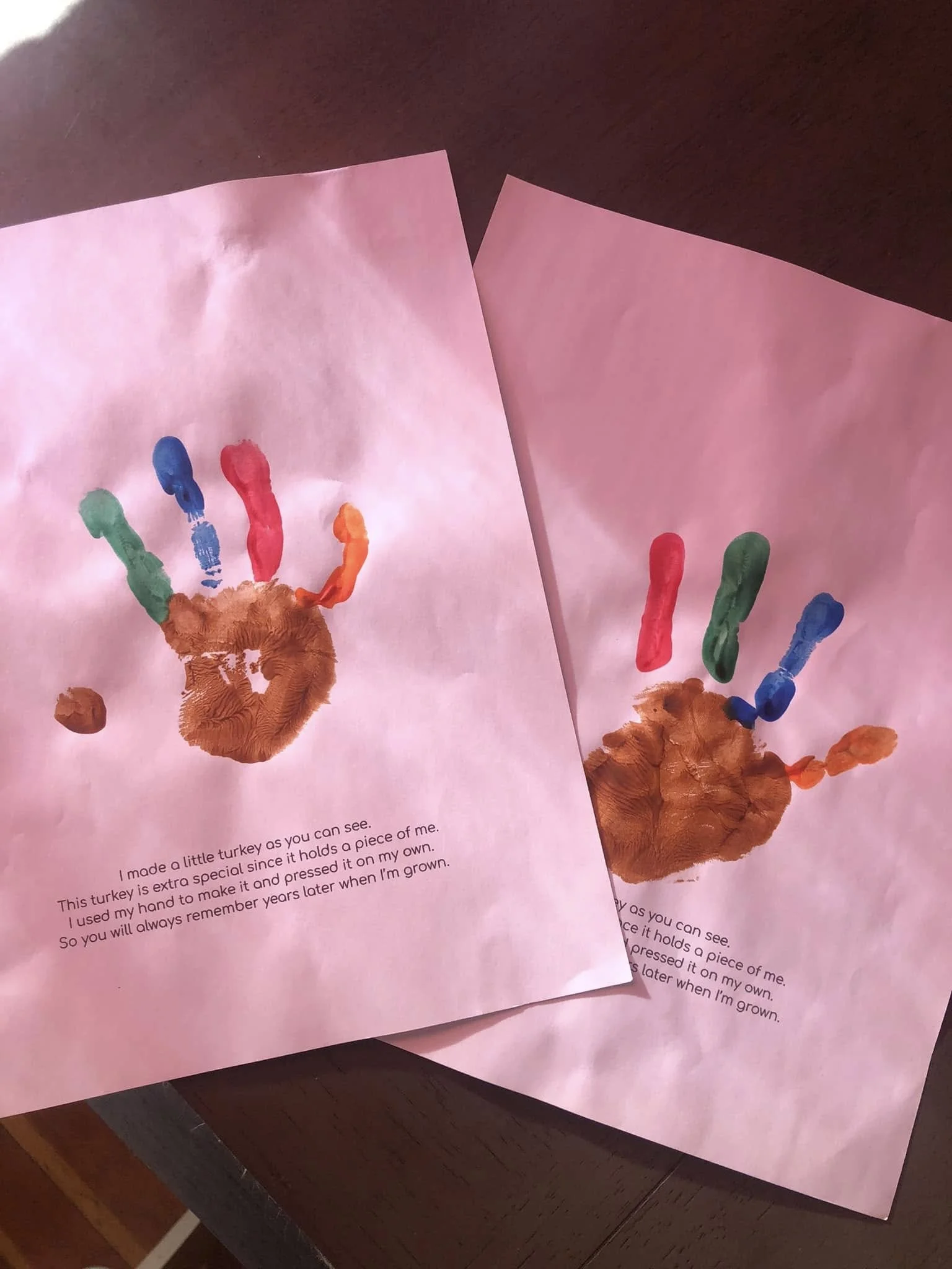 Two pink sheets of paper lying on a wooden surface, each with a handprint made from brown finger paint, with fingers painted in various colors including green, blue, red, orange, and yellow, accompanied by a typed message about making a special piece