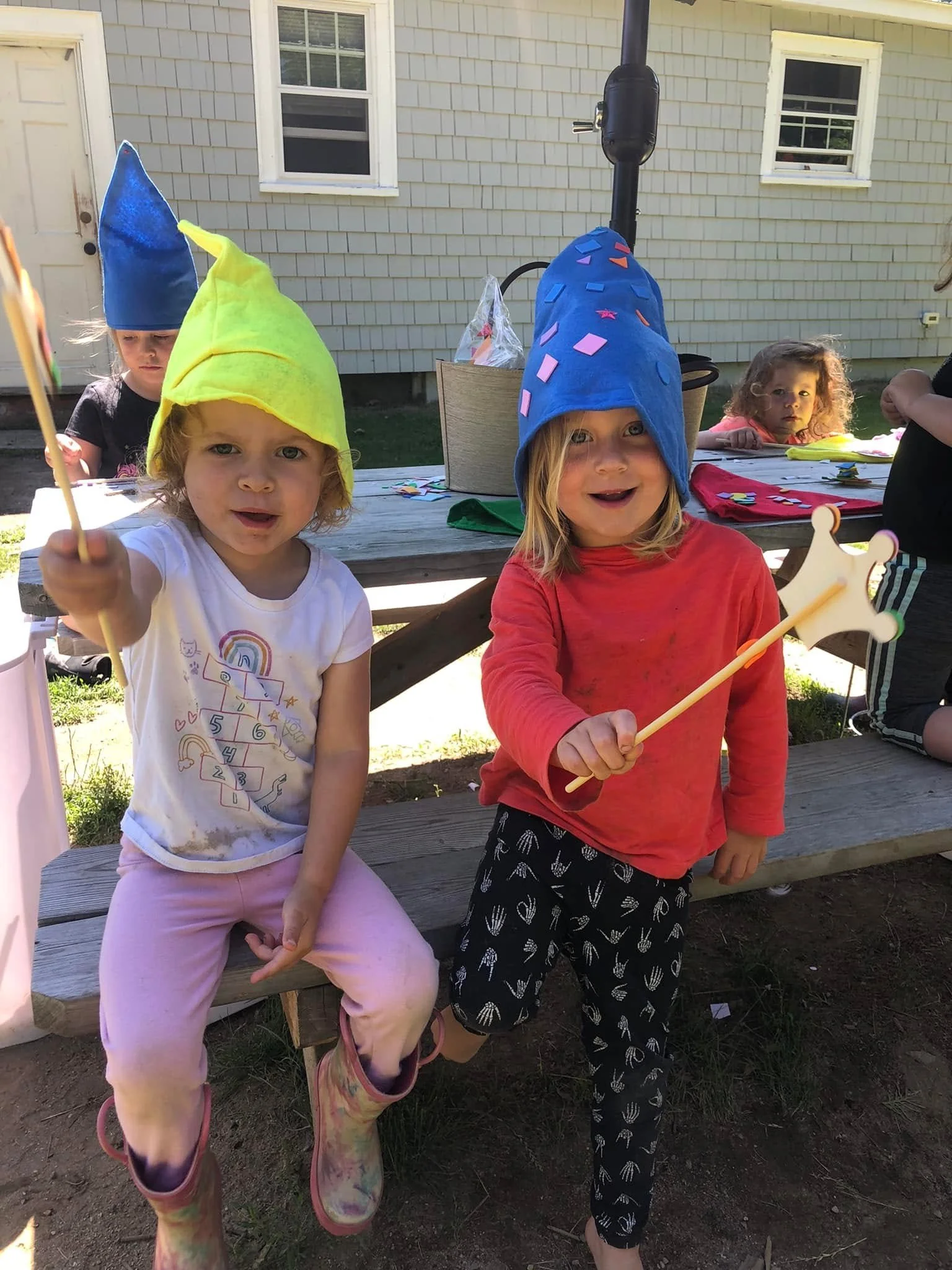 Children wearing colorful wizard hats at a backyard gathering, with a wooden table and house in the background.