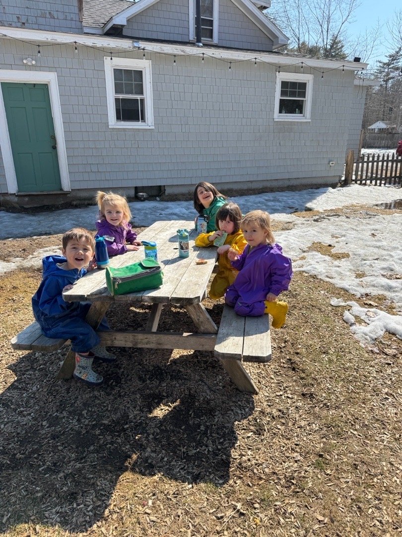 Six children sitting around an outdoor picnic table in a yard with patches of snow, a house with gray siding and white trim in the background, some children wearing colorful rain jackets and boots, eating snacks and smiling.