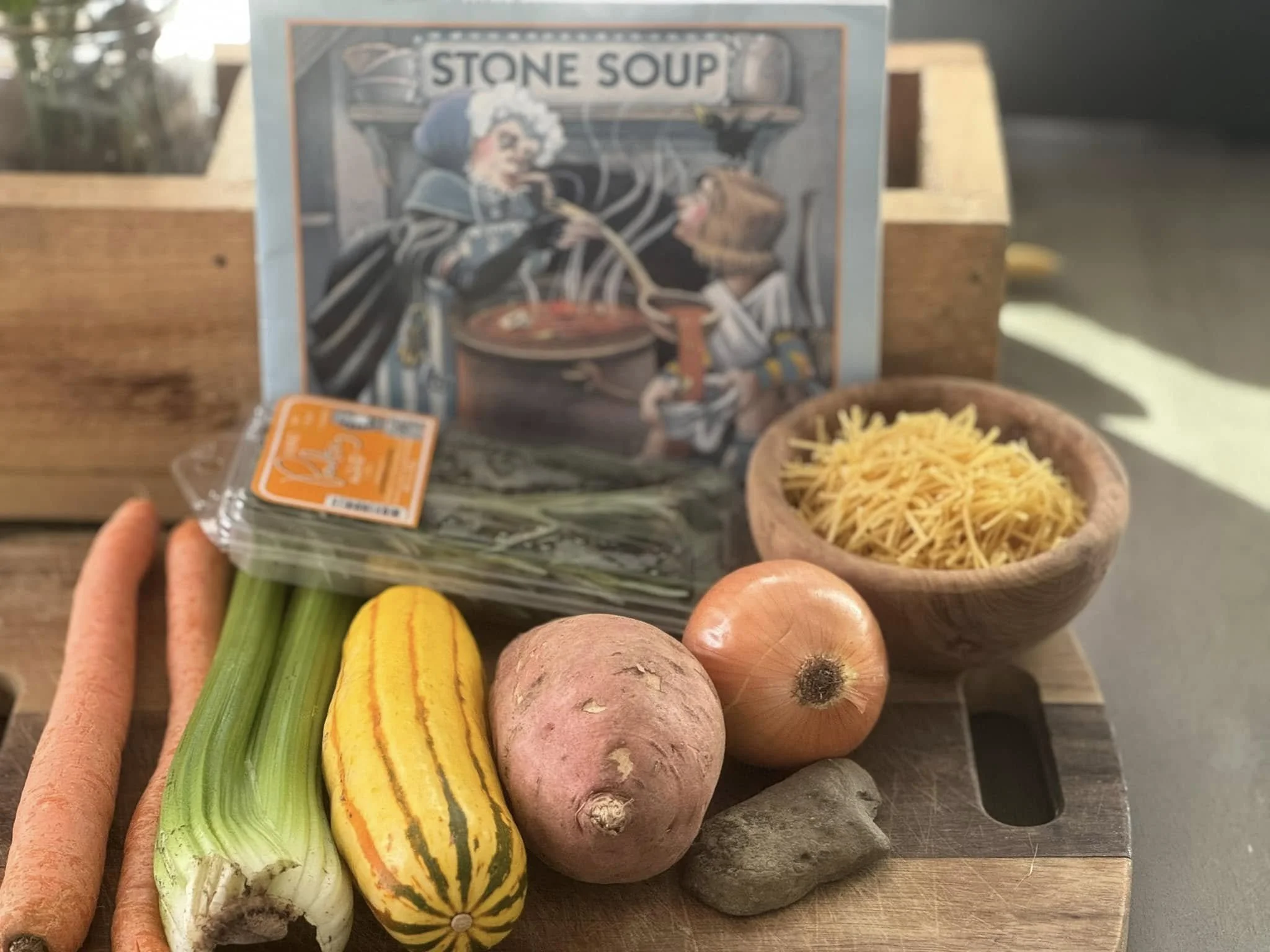 A variety of vegetables including carrots, tomatoes, green onions, yellow squash, sweet potatoes, and onions are displayed on a wooden surface. In the background, there is a box of stone soup and a bowl of shredded cheese.