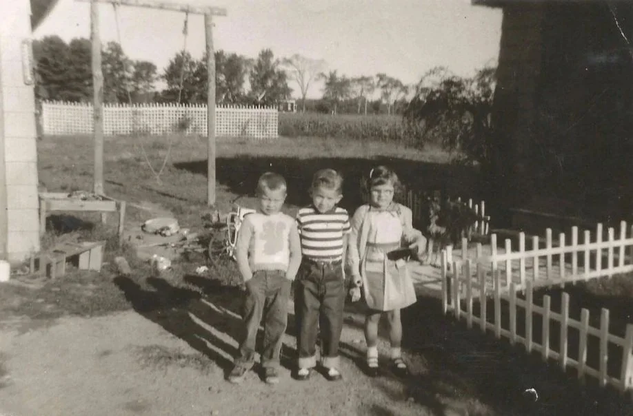Black and white photo of three children standing outdoors in front of a small white fence, with a grassy yard and trees in the background. The children are posing for the photo with the middle child holding something in their hand.