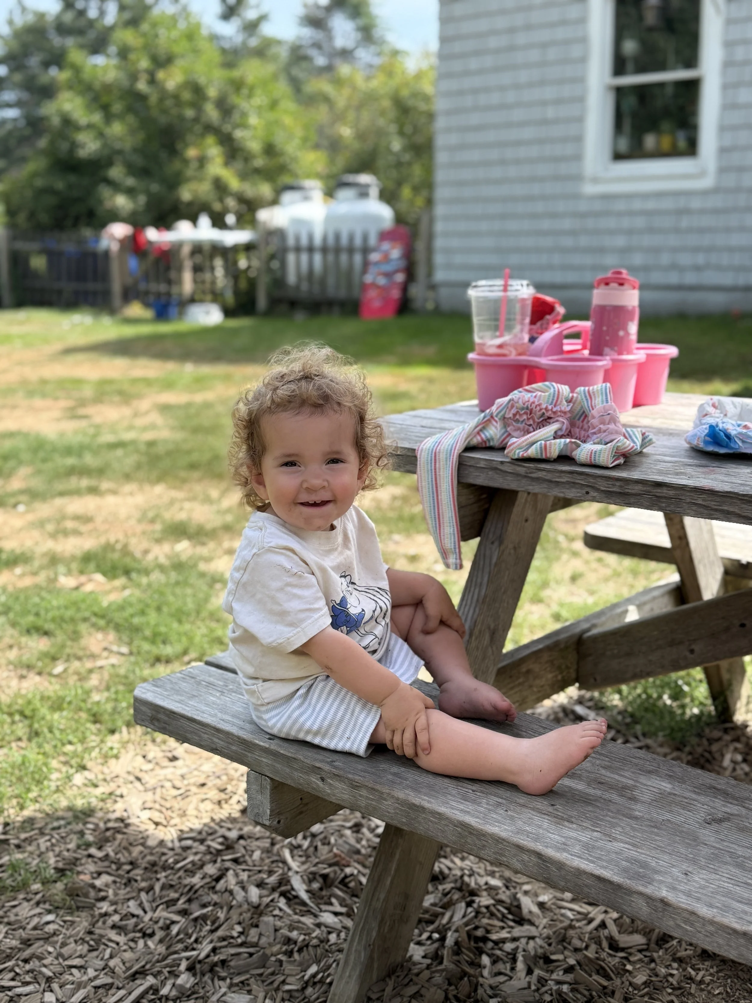 A young child with curly hair and a white shirt sitting on a wooden picnic table outdoors, smiling at the camera. In the background, there are trees, a house with gray siding, and a pink container with various items on the table.