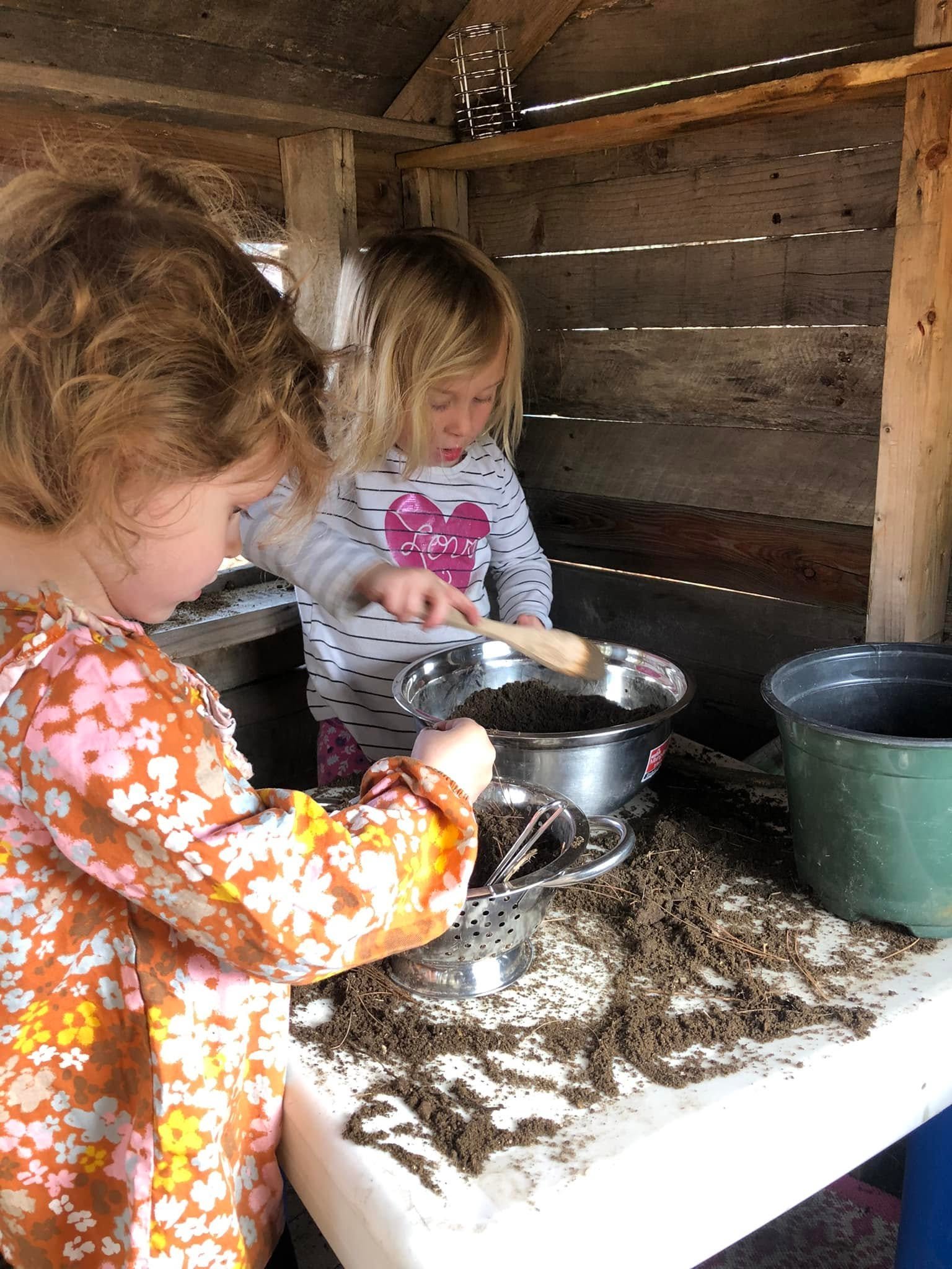 Two girls with curly hair, one wearing an orange patterned shirt and the other wearing a striped shirt with a pink heart, are engaging in an activity involving soil or dirt in a rustic wooden shed. They are scooping dirt into metal containers on a wh