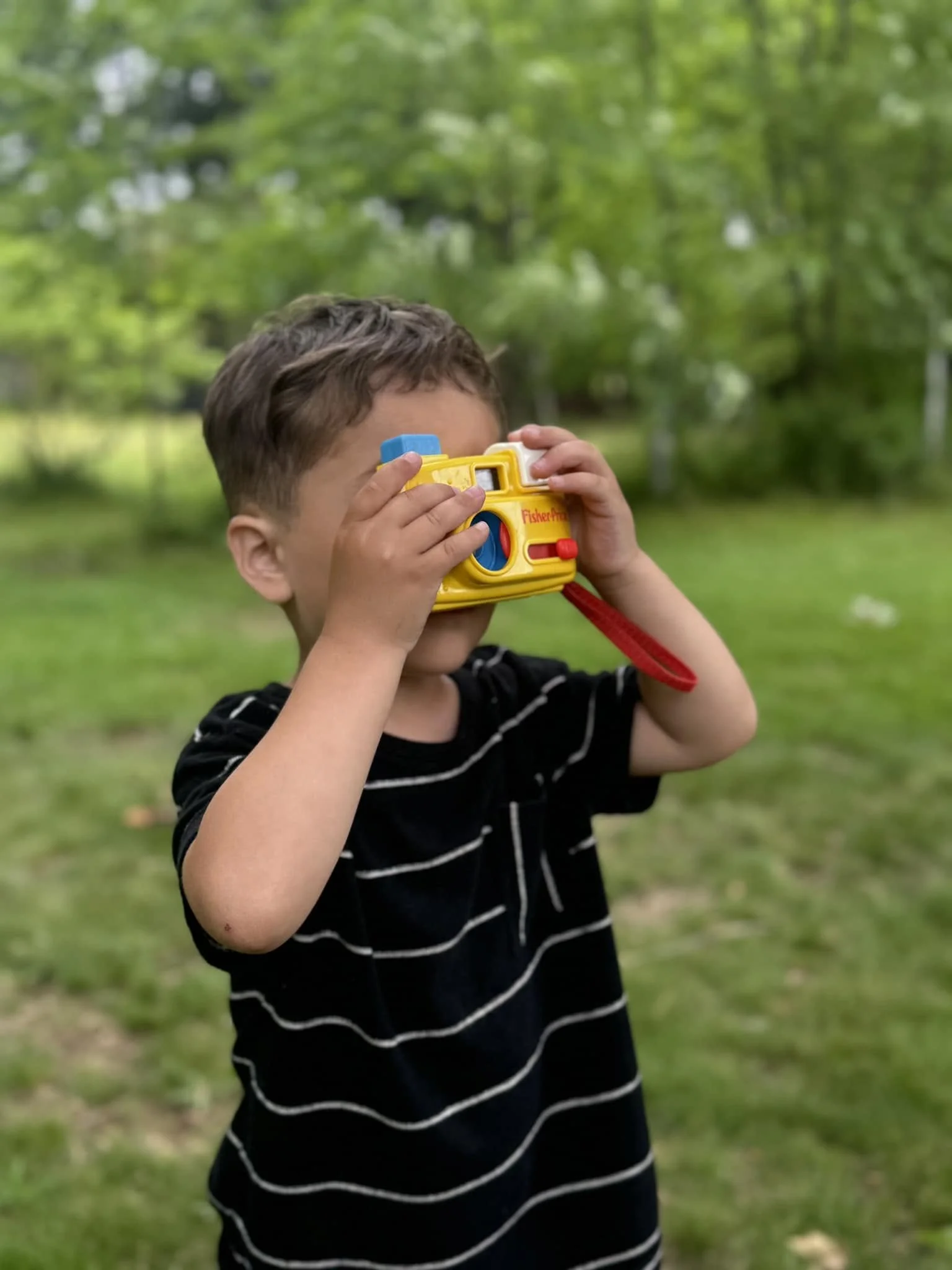 A young boy wearing a black and white striped shirt is holding a colorful Fisher-Price toy camera up to his face outdoors, surrounded by lush green trees and grass.