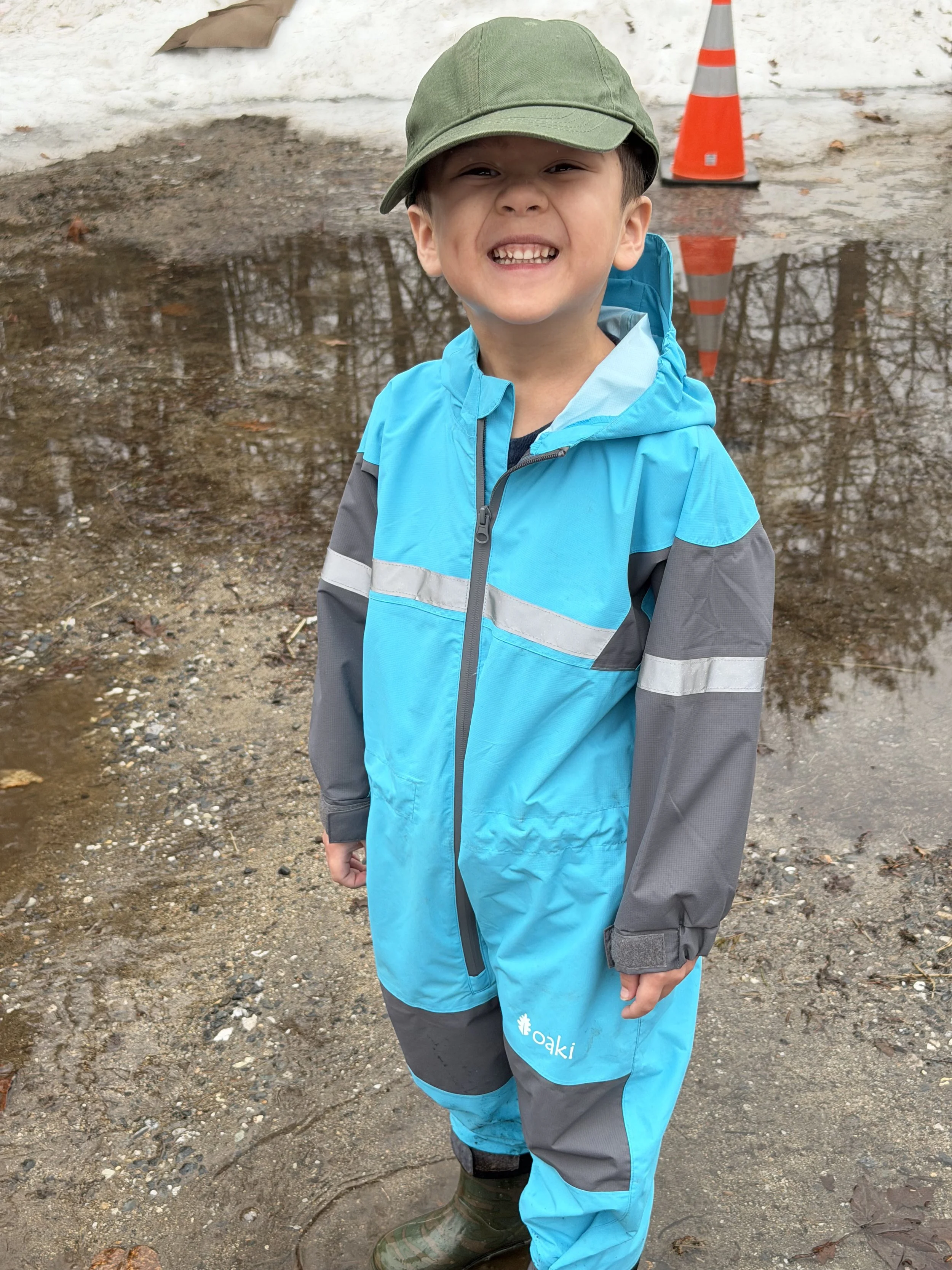 A young boy wearing a green cap, a blue and gray waterproof jacket, and green waterproof boots, standing outdoors in a muddy area with puddles and leafless trees reflected in the water.