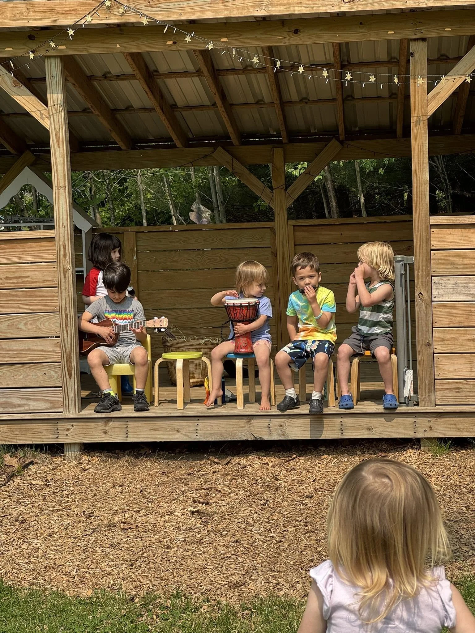 A group of children sitting on chairs under a wooden patio, with some playing musical instruments and others talking. A girl with blonde hair is in the foreground, watching the children on the patio.