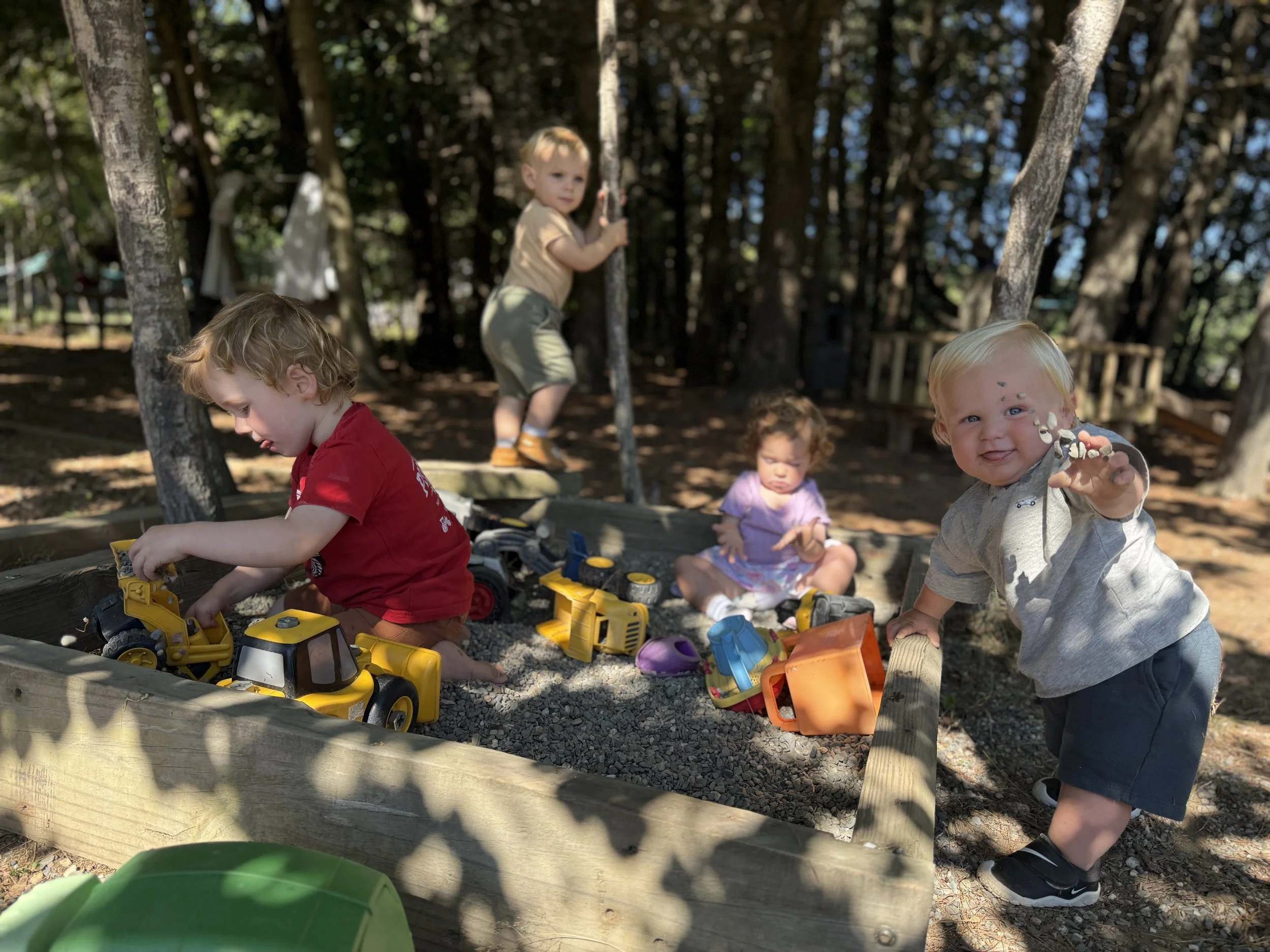 A group of young children playing in a sandbox outdoors surrounded by trees, with some children digging and playing with toy trucks, and one child smiling and holding a handful of sand.