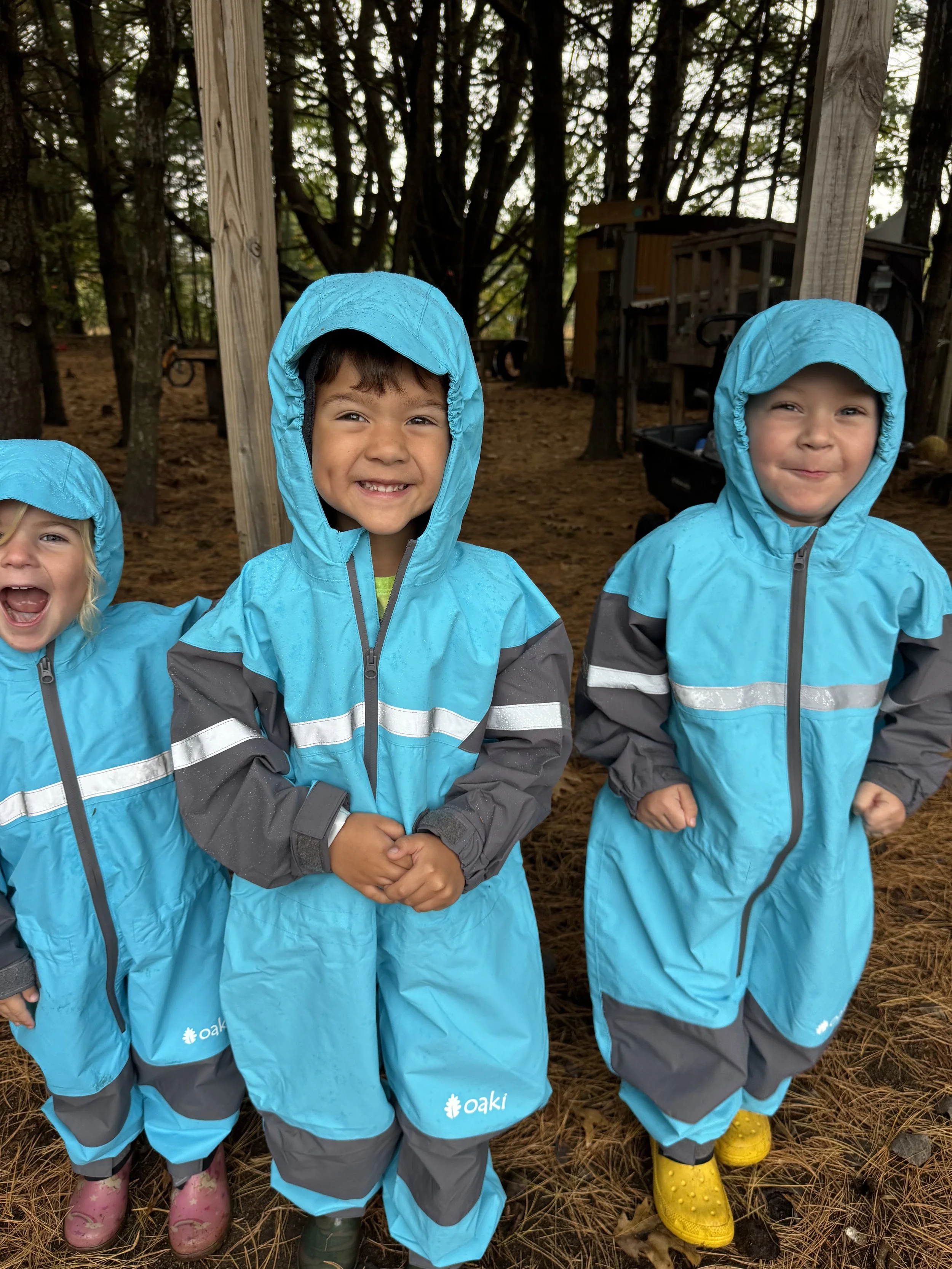 Three young children wearing blue rain jackets and colorful boots standing outdoors on a forest floor with trees and wooden structures in the background.