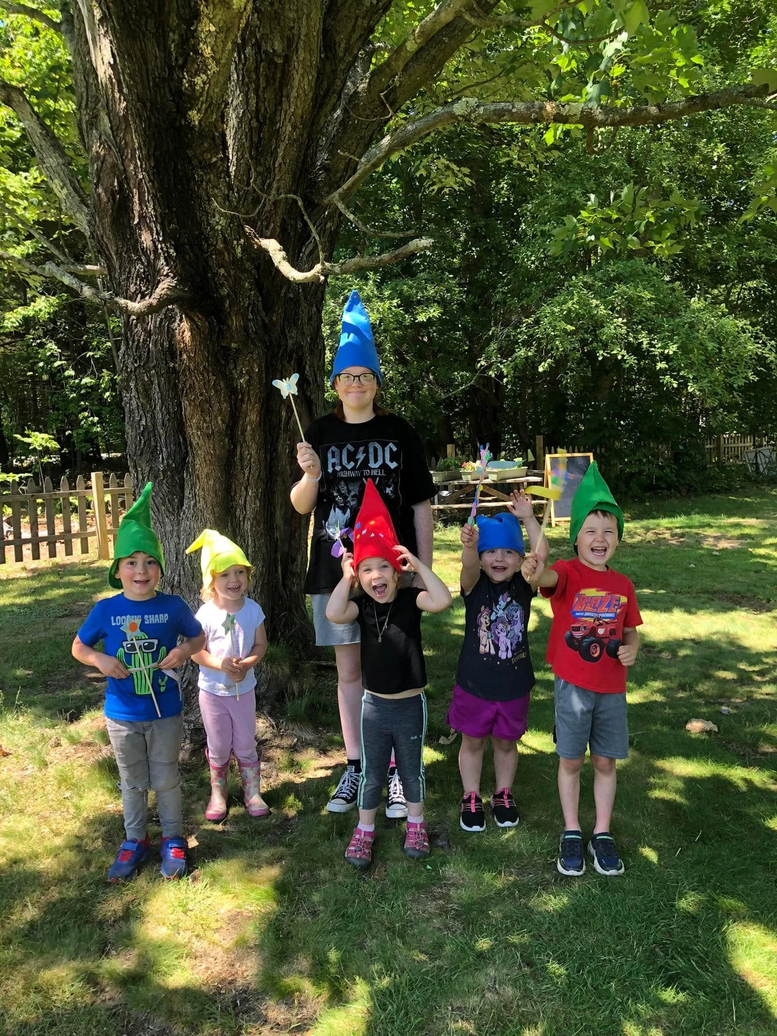 Group of children with party hats and fairy wands outdoors near a large tree, with a woman standing behind them, smiling.