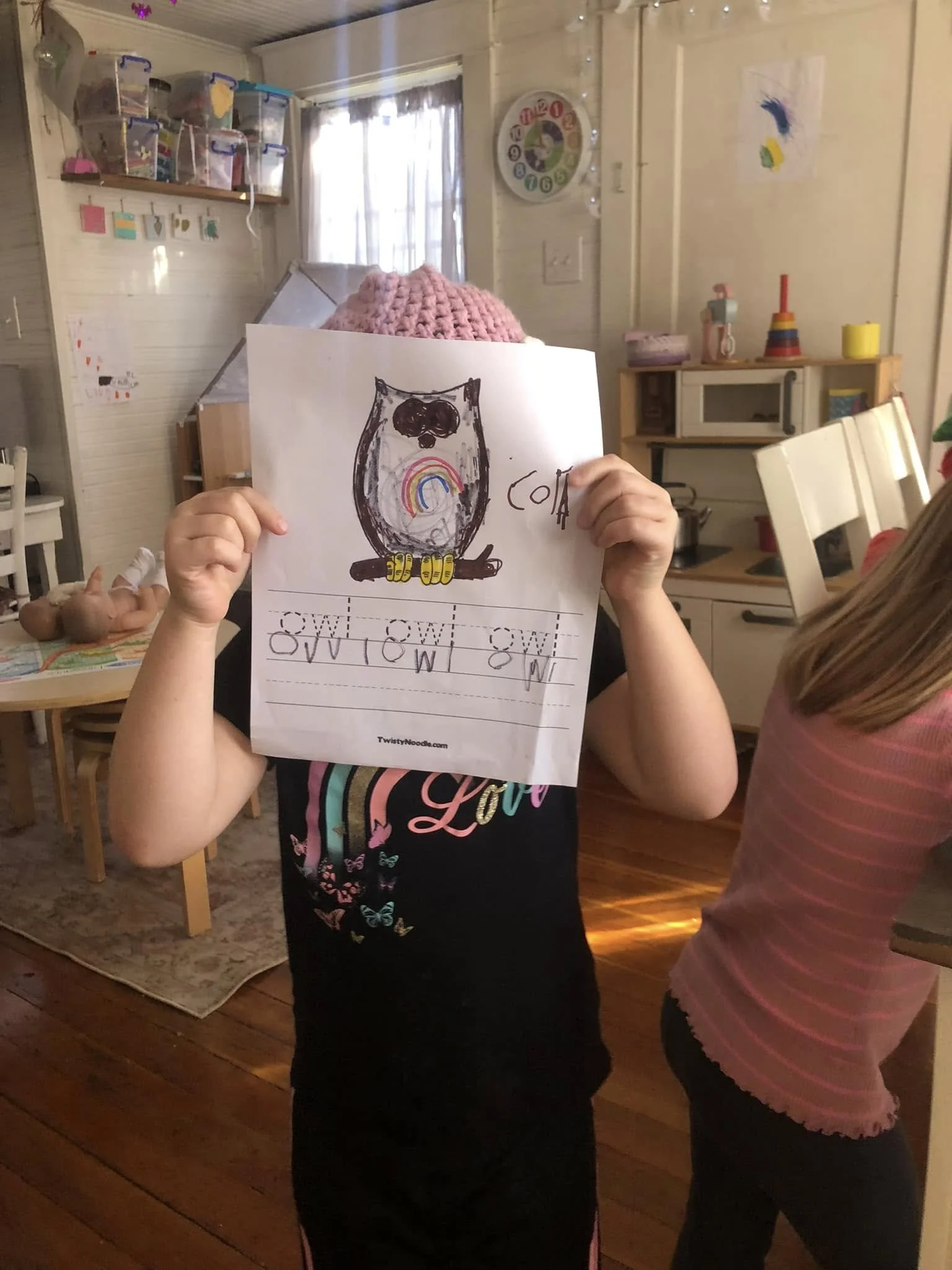 Child holding a coloring sheet with an owl illustration, wearing a pink knit hat, in a cozy living room with wooden floors, kitchen cabinets, and children's artwork on the wall.