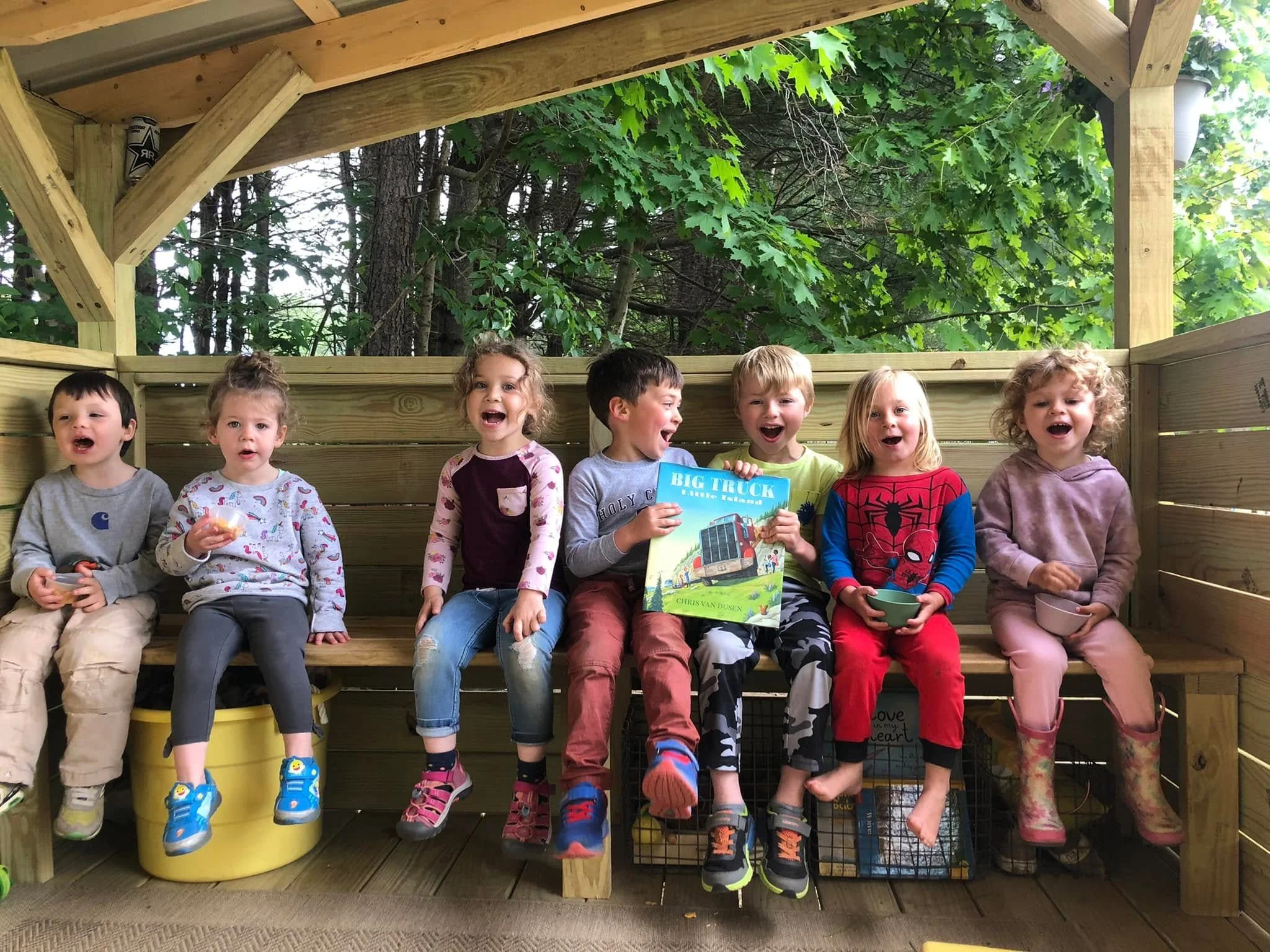 A group of seven children sitting on a wooden deck, smiling and holding toys, with trees and greenery in the background.