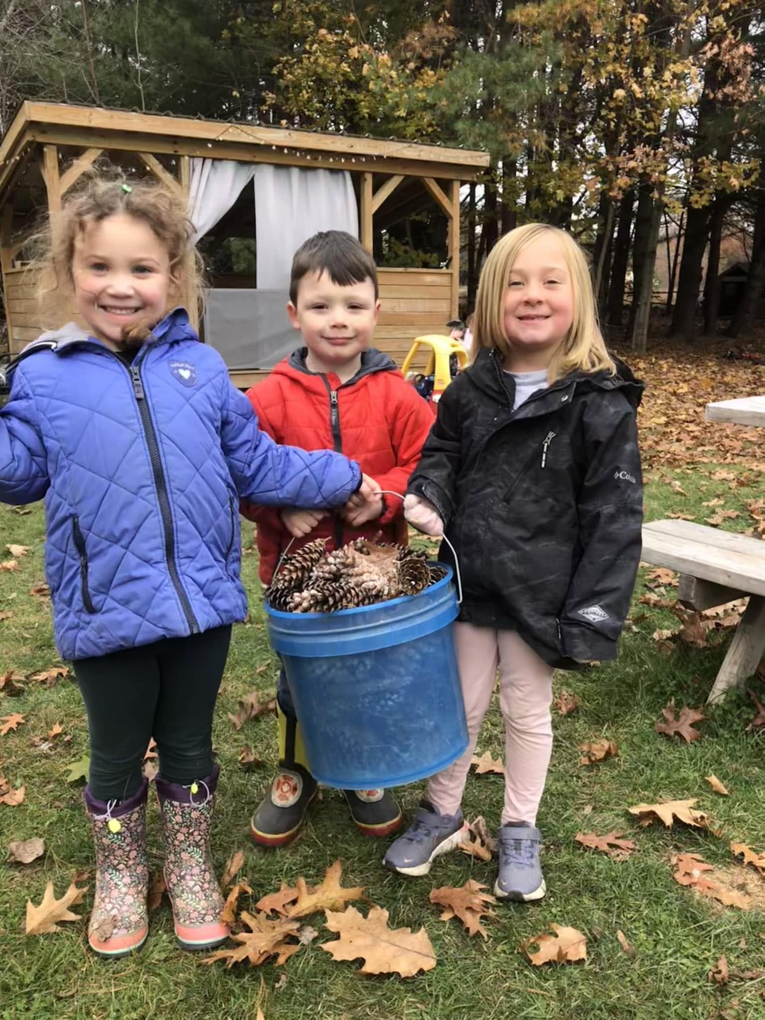 Three children standing outdoors on fall day, holding a bucket filled with pinecones, with a small wooden playhouse and trees with autumn leaves in background.