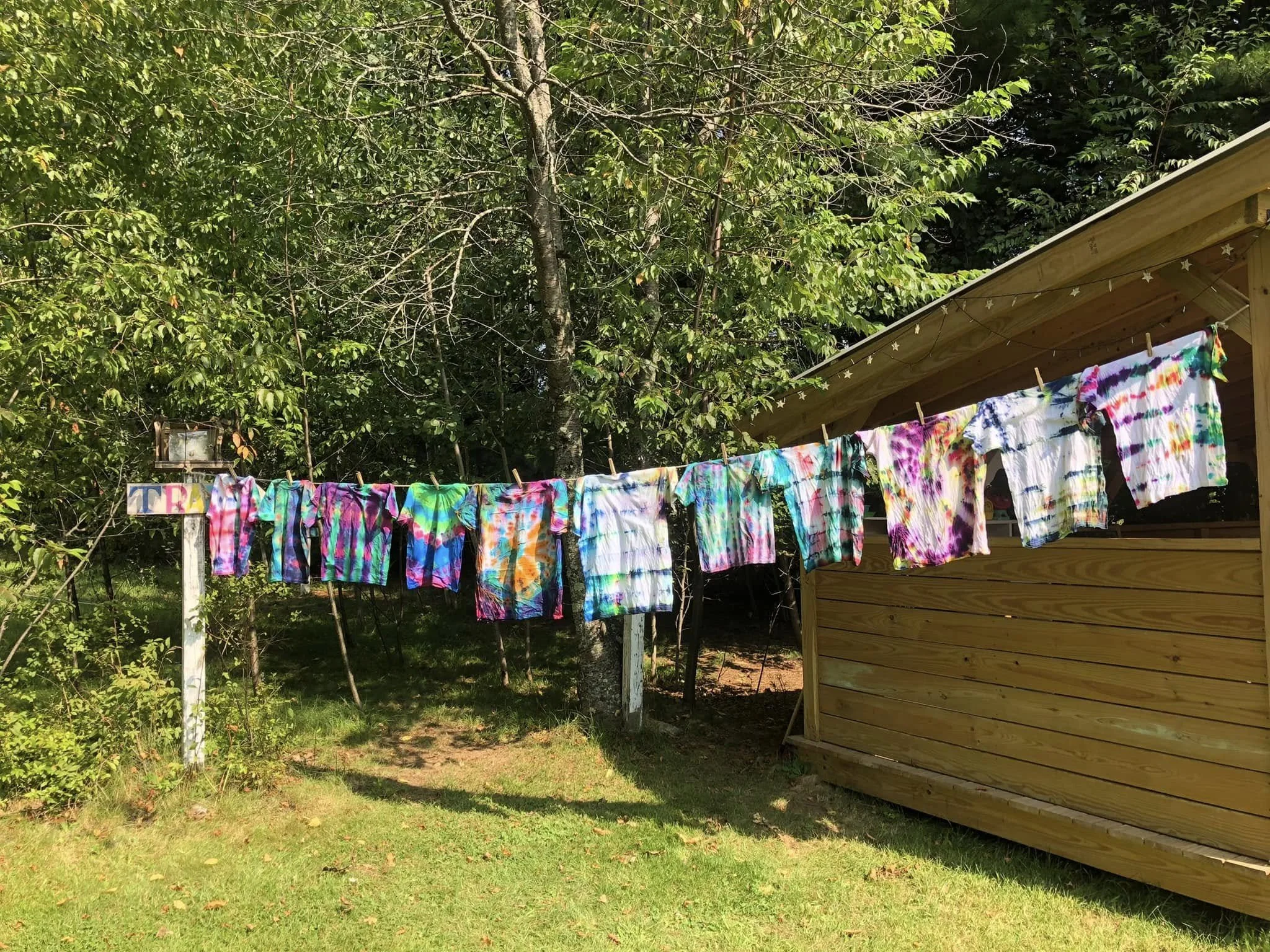 Colorful tie-dye shirts hanging on a clothesline outdoors, with trees and a wooden shed in the background on a sunny day.