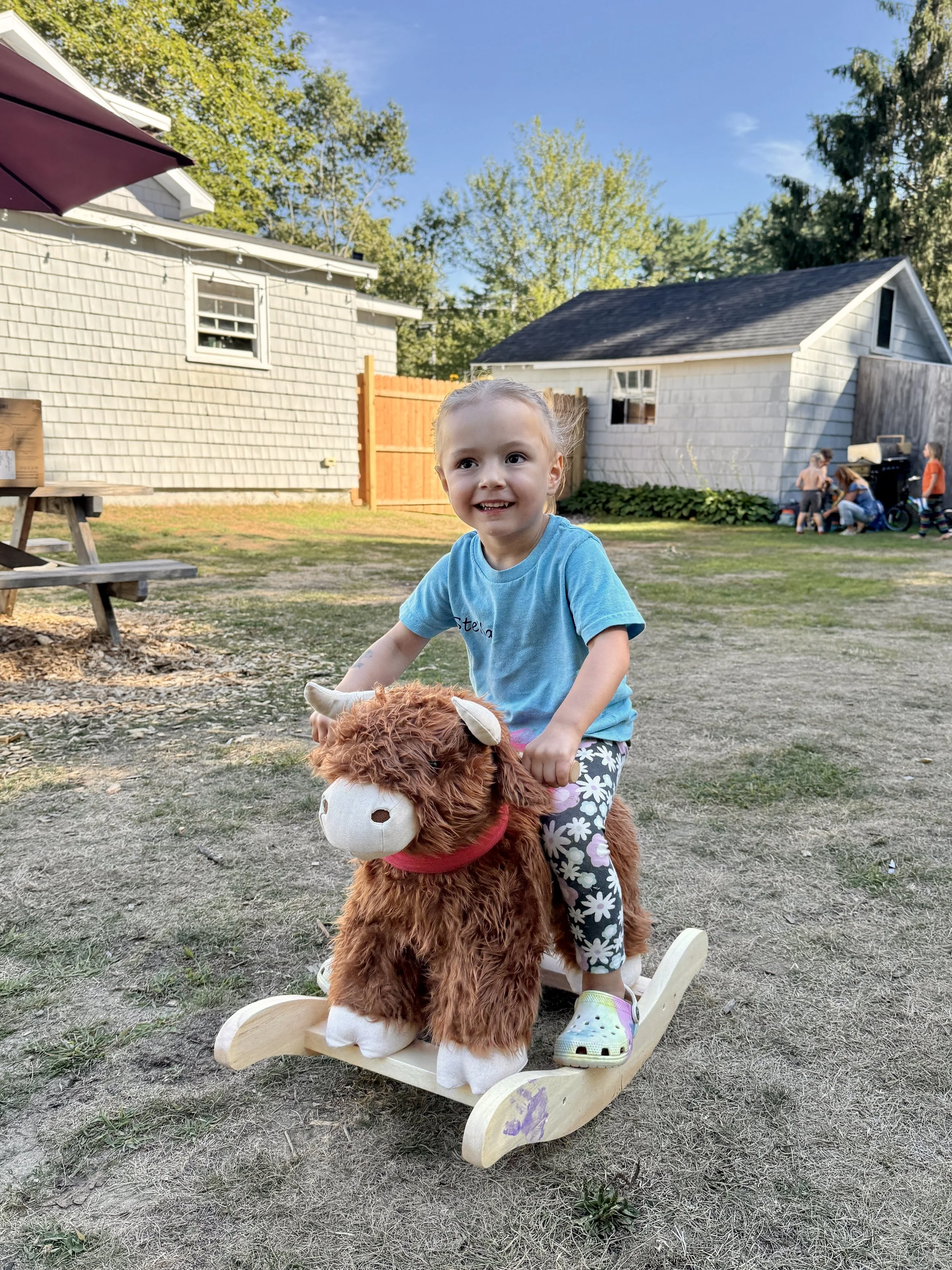 Young girl riding a wooden rocking horse outdoors in a backyard with two small white buildings, one with shingle siding and the other with lap siding, and a group of children in the distance. The girl is wearing a blue T-shirt and floral pants, smili