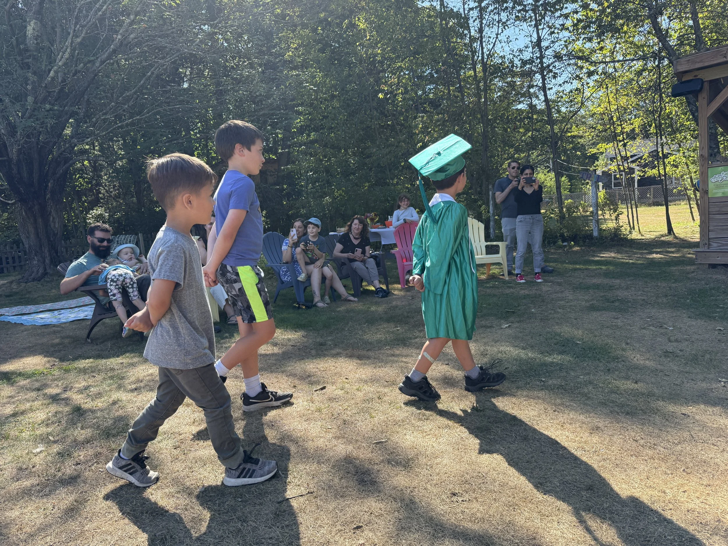 Children participating in an outdoor graduation ceremony, with some seated and others standing, and an adult taking photos amidst trees and chairs.